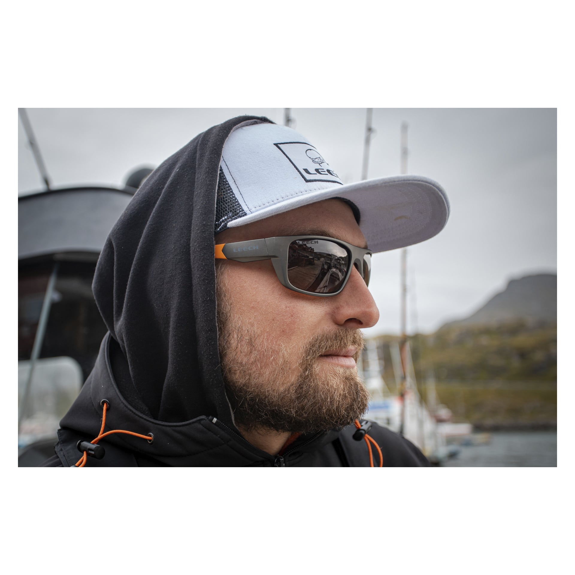 En skäggig person, klädd i svart hoodie, grå keps och mörka solglasögon med kopparfärgade Moonstone Orange-linser från Leech Fishing Glasses, står vid vattnet där båtar ligger förtöjda. Bakom dem breder sig ett stenigt landskap under en molnig himmel.