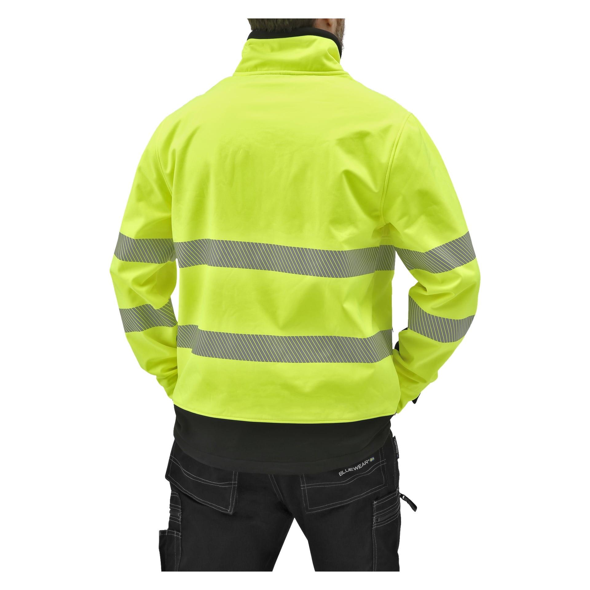 Sedd bakifrån står en person med händerna på höfterna, klädd i en klargul Bluewear Hi-Vis softshell-jacka Toronto med långärmat och reflekterande horisontella ränder, tillsammans med svarta byxor.