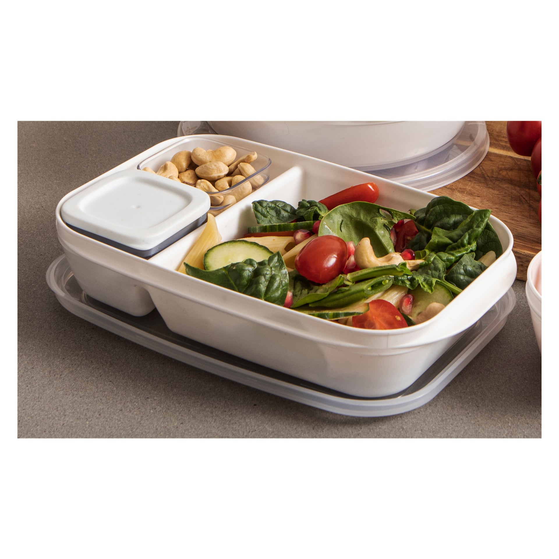 En Menuett Lunchbox med delade sektioner innehåller en spenatsallad med körsbärstomater, cashewnötter, granatäppelkärnor, gurkskivor, en portion pasta och en liten kopp vit dressing på en grå bänkskiva.