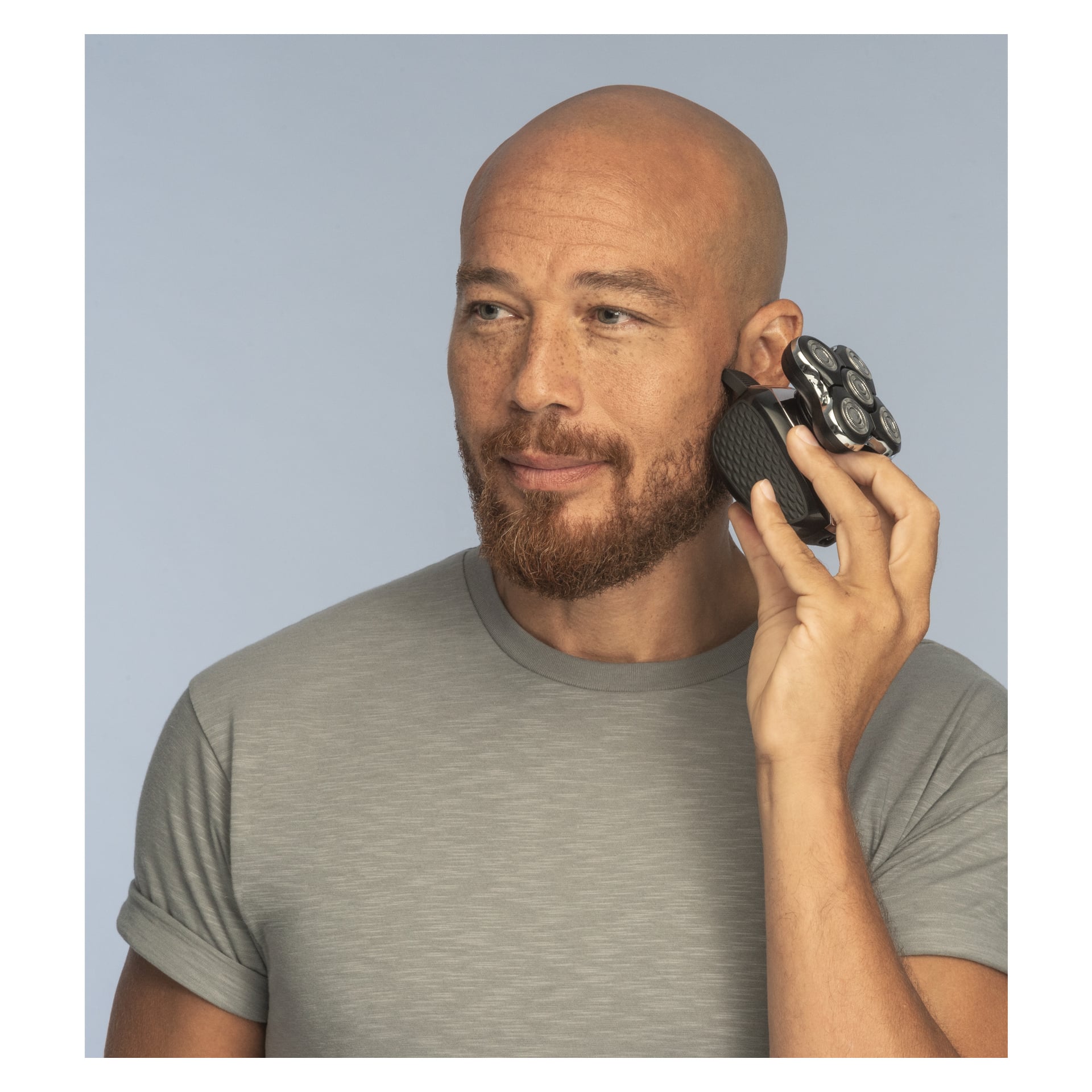 En skallig man med skägg, klädd i en ljusgrå t-shirt, håller Remington Cordless rakhyvel för huvudet Ultimate Series XR1500 med sina runda rattar upp till örat framför en enkel ljusblå bakgrund.