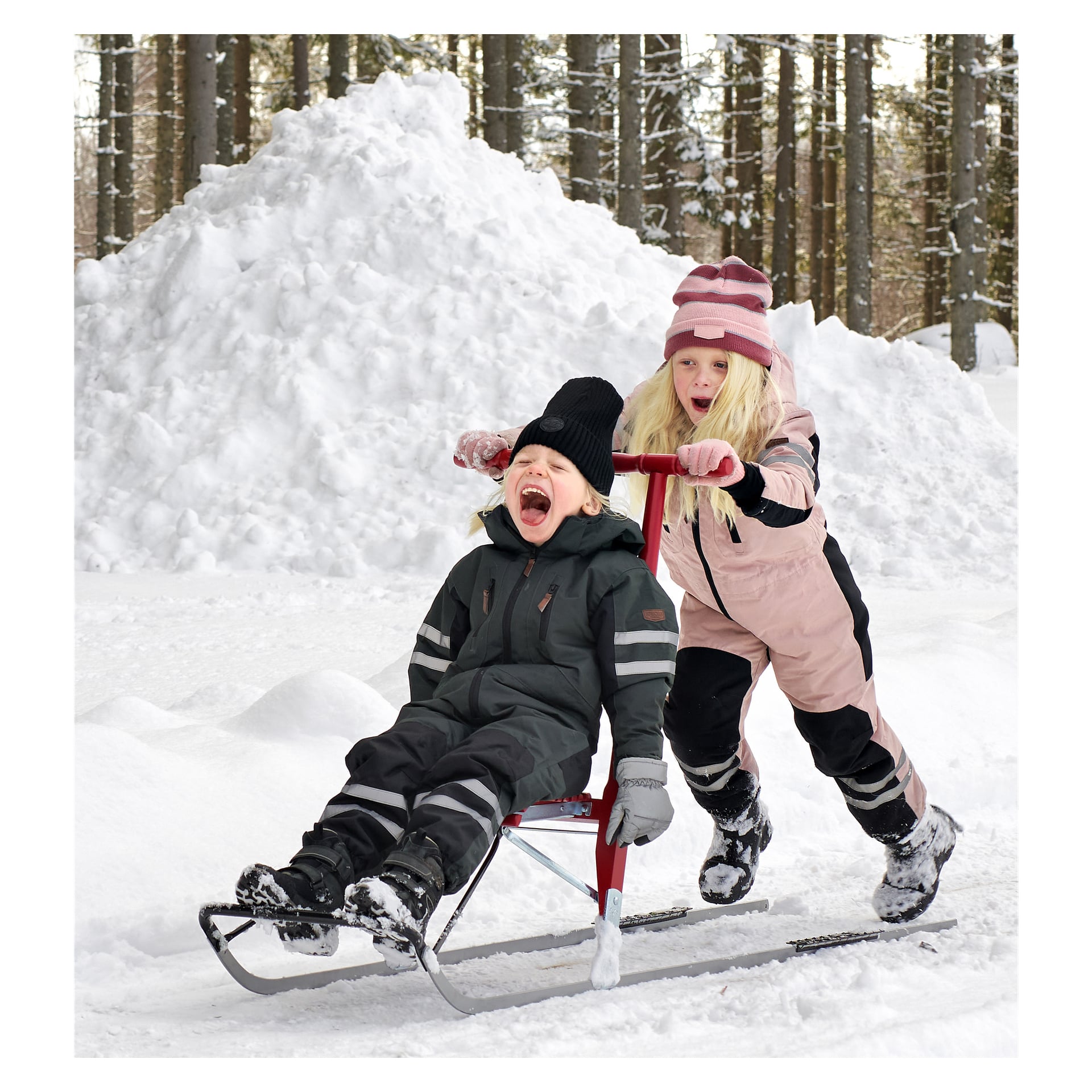 Två barn leker i snön. Barnet i svart skriker medan det åker en Gustafvsivers Scooter Sputte-pulka, och den som är klädd i rosa knuffar bakifrån. I bakgrunden finns snö och träd.