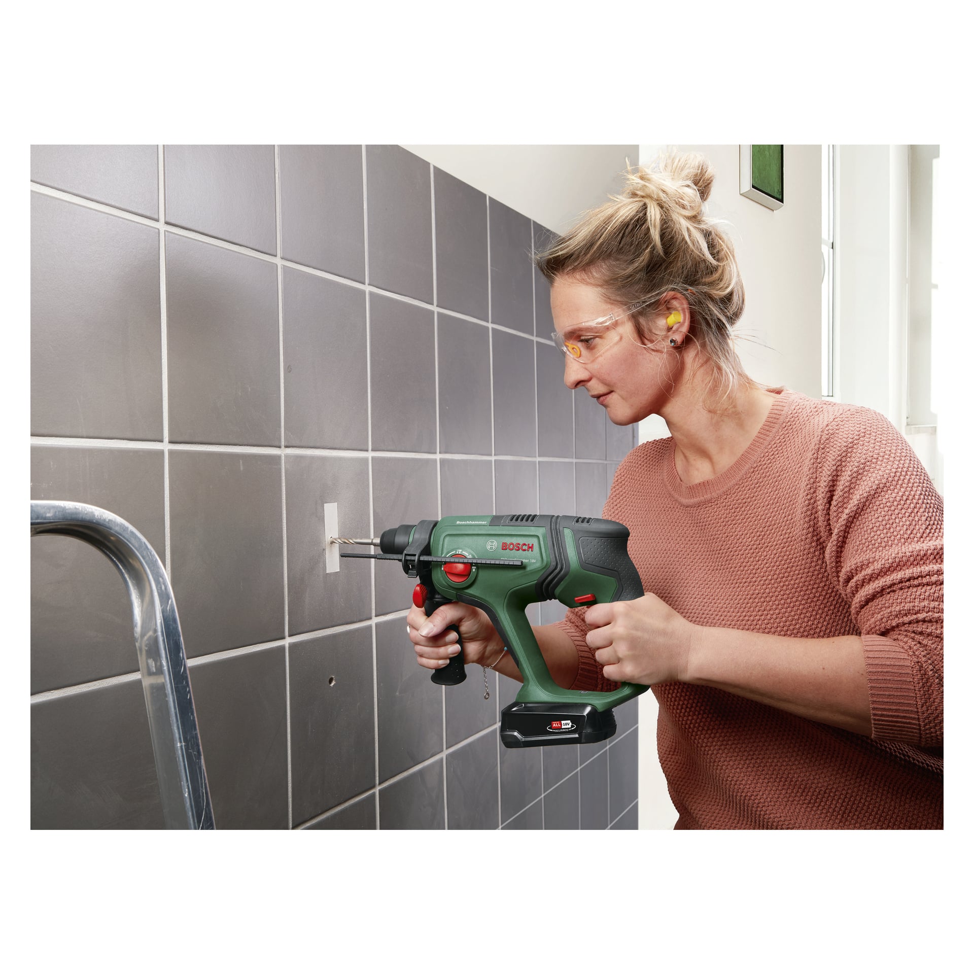 En kvinna, utrustad med skyddsglasögon och öronproppar, använder en Bosch Power 4 All UniversalHammer 18V sladdlös borrmaskin för att borra i en kaklad vägg. En del av en metallstege syns i förgrunden.