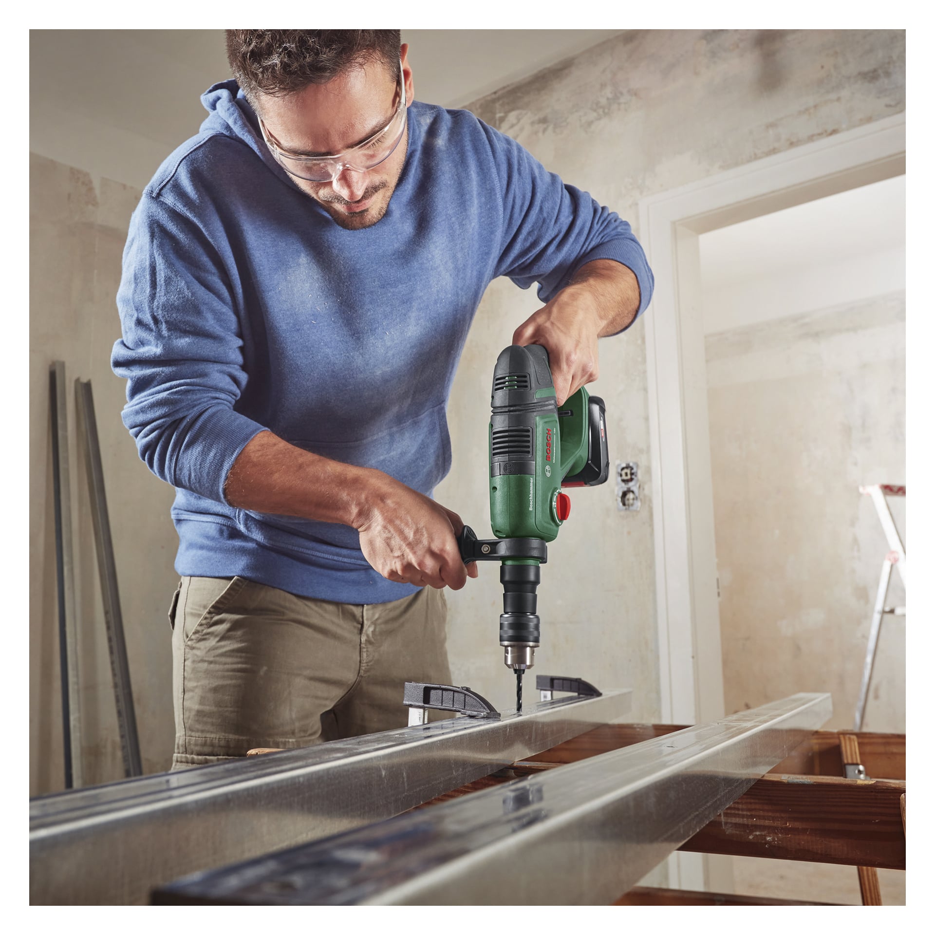 En man med skyddsglasögon och blå hoodie använder en Bosch Power 4 All UniversalHammer 18V sladdlös hammarborrmaskin och borrar i en metallbalk som står på en arbetsbänk i ett delvis renoverat rum.
