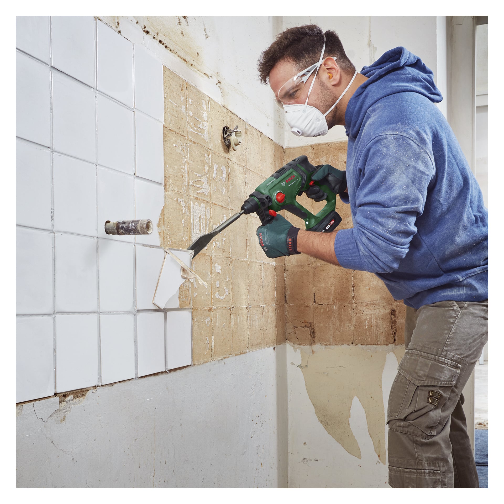 Iklädd en blå hoodie, skyddsglasögon och dammmask använder en arbetare Bosch Power 4 All UniversalHammer 18V batteridriven hammarborr för att ta bort vita kakel från en köksstänkskydd.