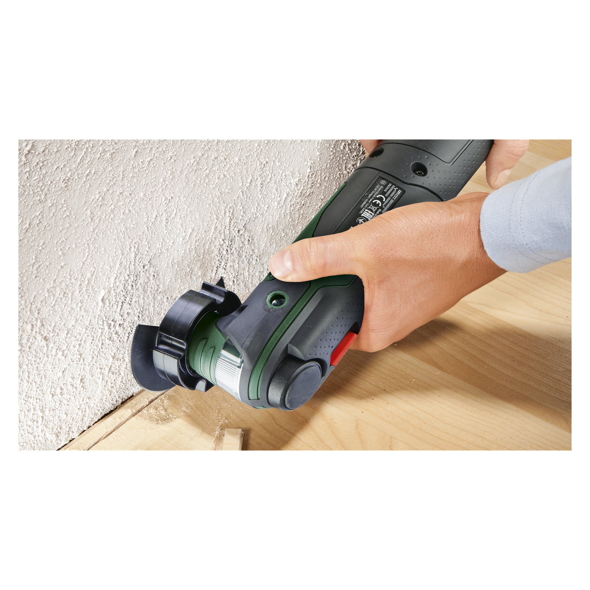Man greppar hårt Bosch Power 4 All Multi-tool AdvancedMulti 18 V med dess gröna och svarta handtag och använder den för att arbeta på kanten av ett trägolv intill en vit vägg med textur.