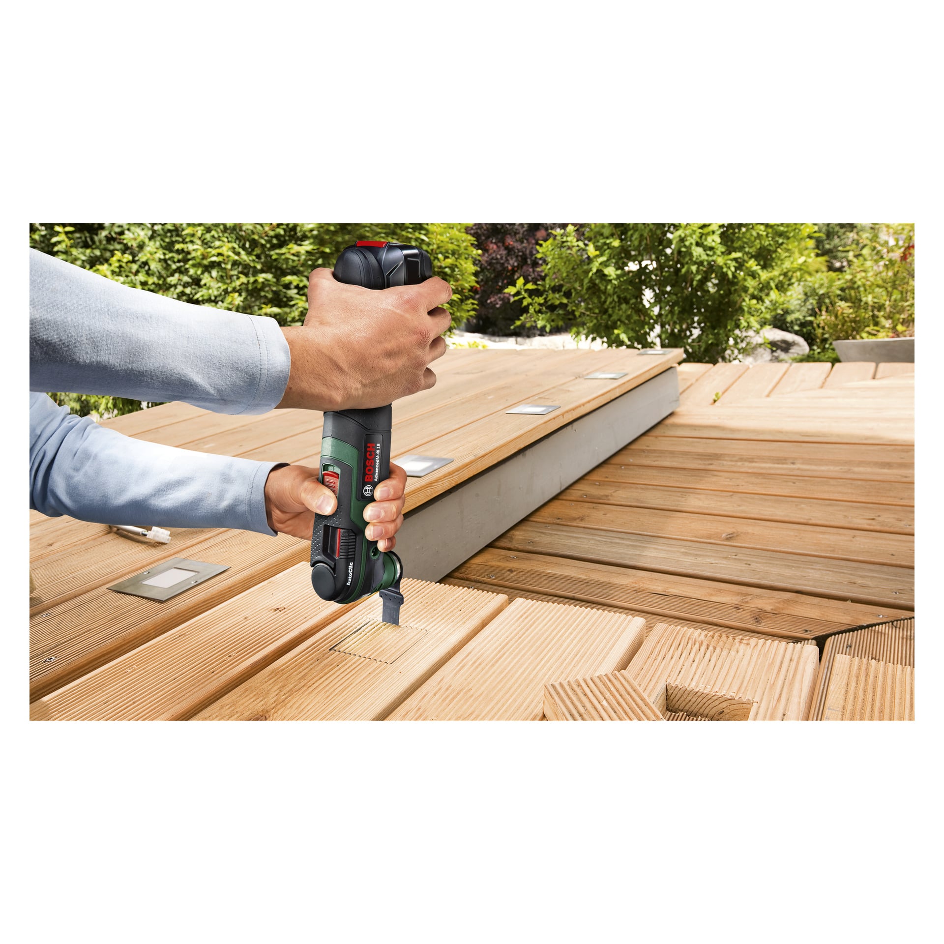 Man använder Bosch Power 4 All Multi-tool AdvancedMulti 18 V för att skära ett rektangulärt hål i en träterrass utomhus, och håller verktyget säkert med båda händerna medan solljuset lyser upp arbetsytan.
