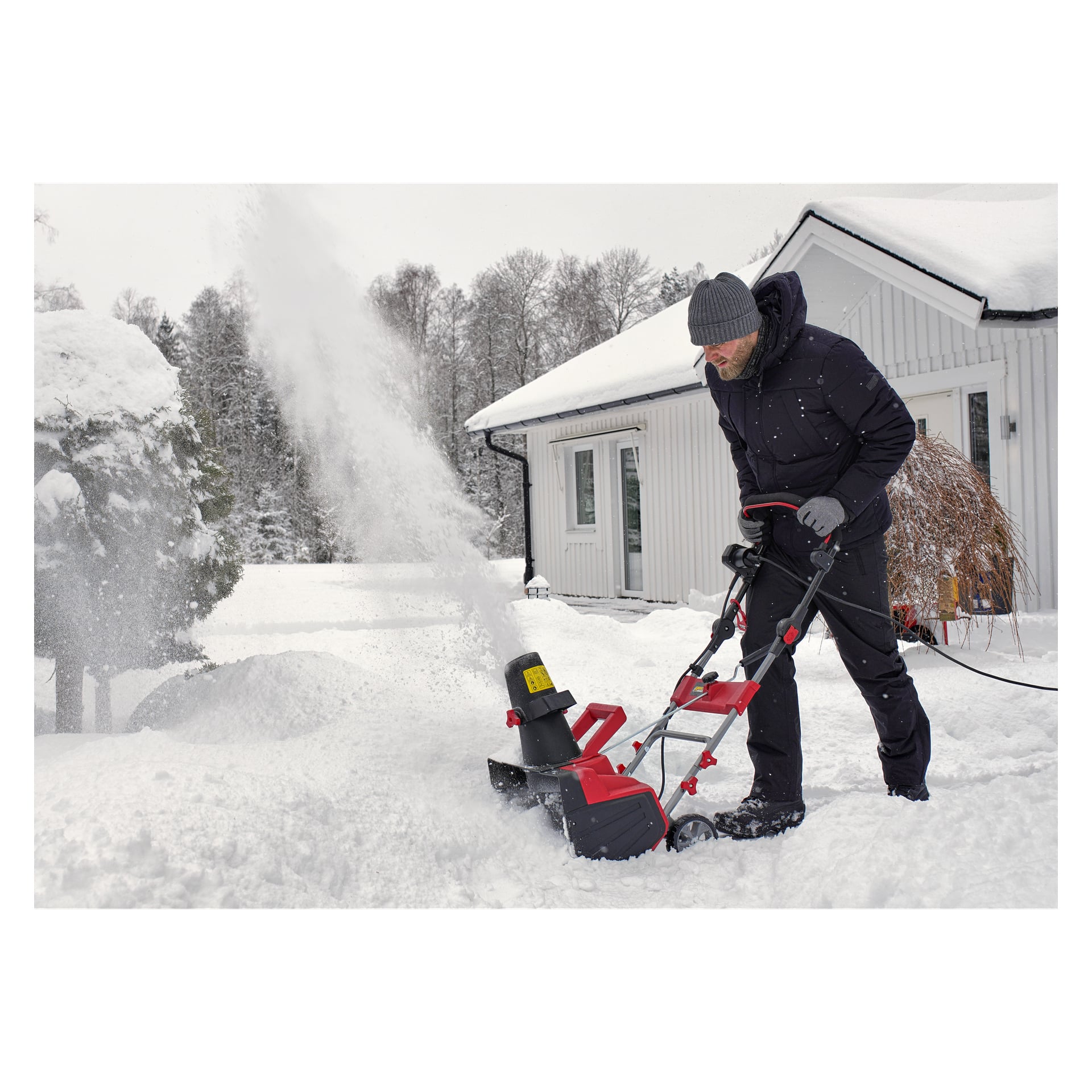 I vinterklær bruker man en elektrisk snøfreser fra Meec Tools for å fjerne snø fra en innkjørsel nær et hvitt hus. Snø flyr gjennom luften mens nakne trær står i bakgrunnen.