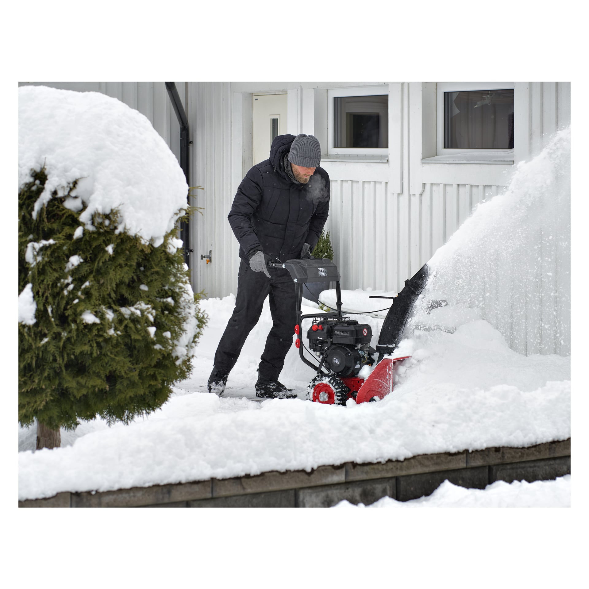 En person i vinterkläder använder en Meec Tools snöslunga för att ta bort snö från en uppfart bredvid ett hus, medan snön sprutar från maskinen och ett litet träd står i förgrunden.