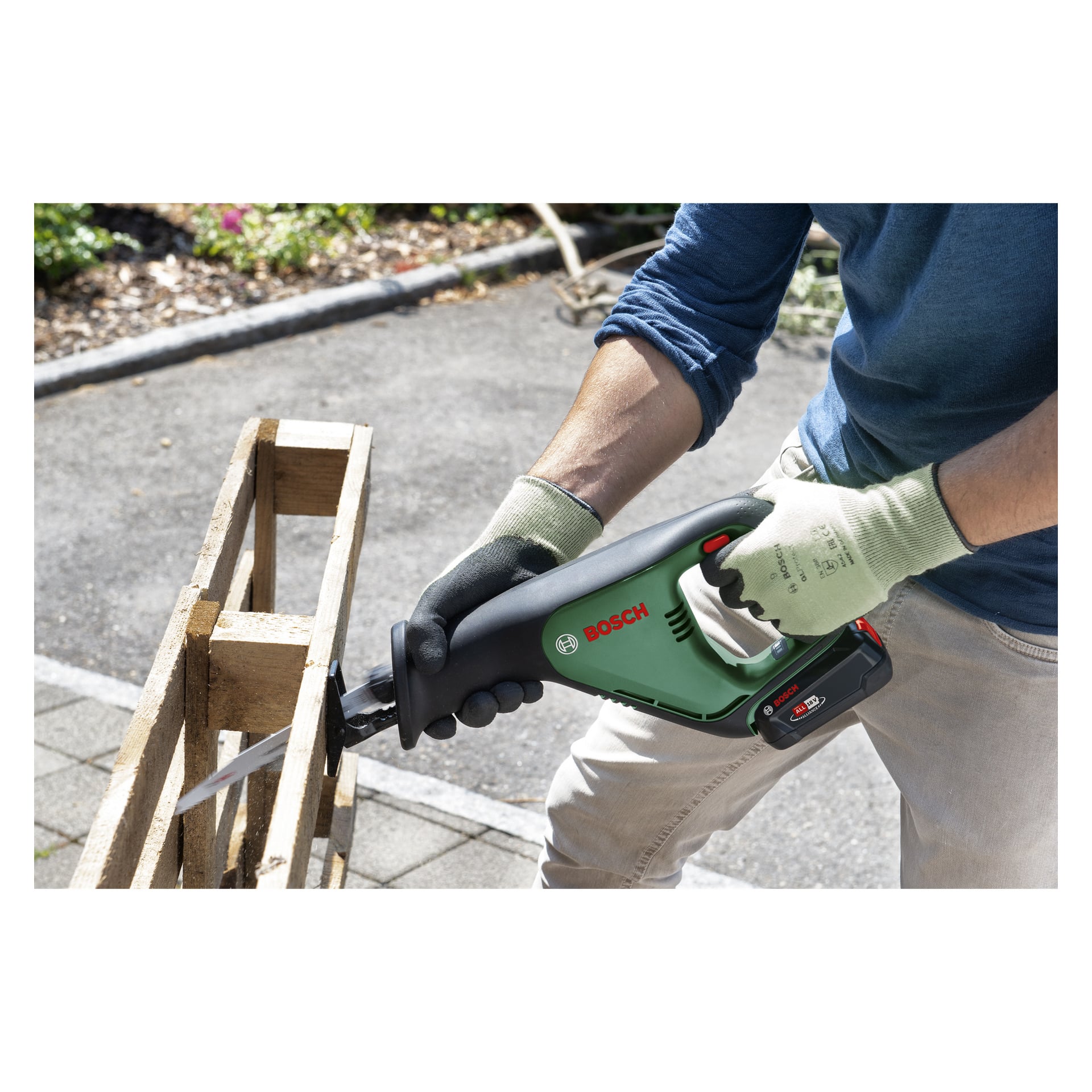 En person med handske använder en Bosch Power 4 All AdvancedRecip 18V batteridriven bajonsåg för att såga igenom en träpall på en stenlagd yta utanför, med grön vegetation i bakgrunden.