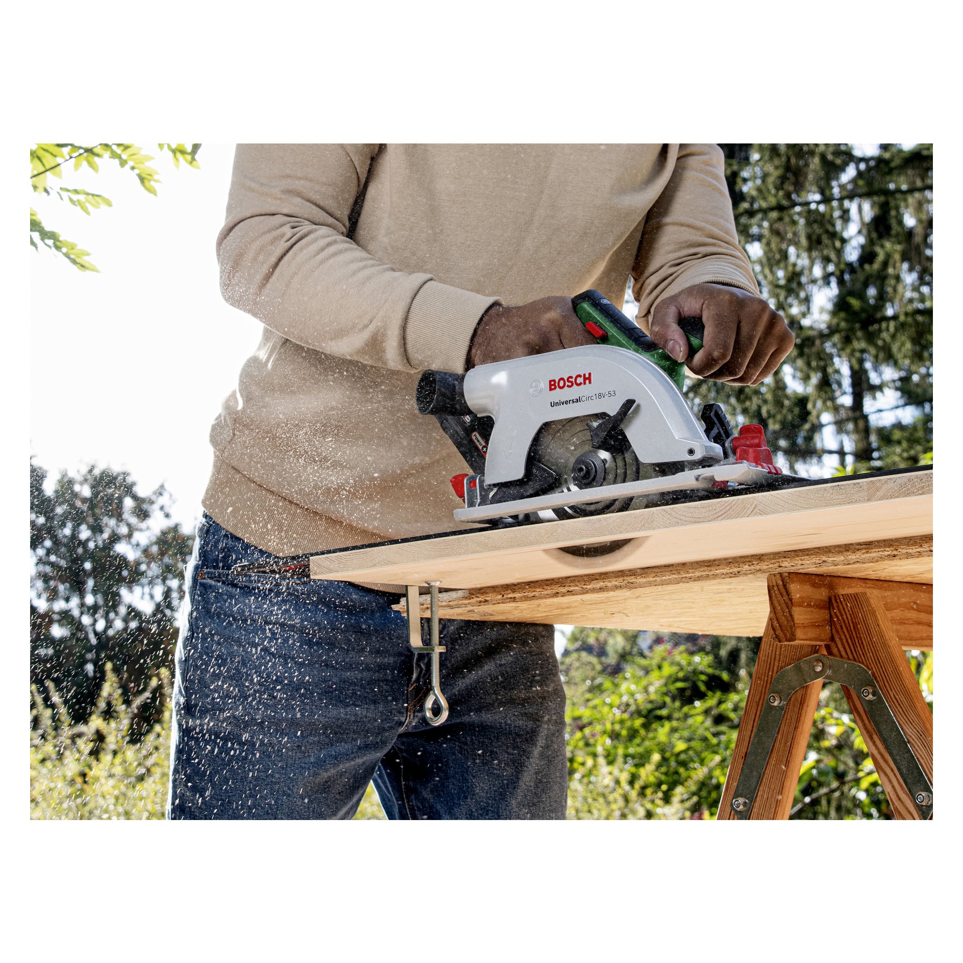 Någon använder en Bosch Power 4 All UniversalCirc 18V-53 sladdlös cirkelsåg för att trimma en träplåt fastsatt på en såghäst, med trädamm som flyger utomhus. Endast personens händer och bål syns i scenen.