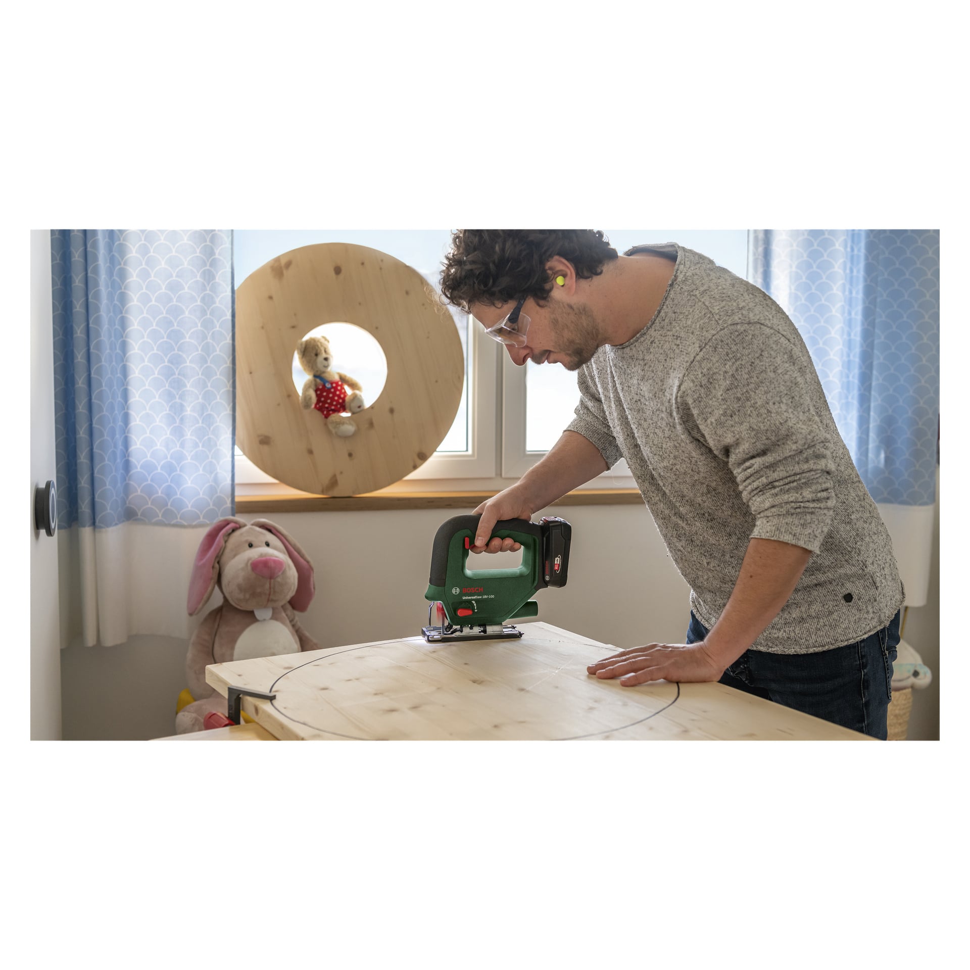 En person, utrustad med skyddsglasögon och öronproppar, använder en Bosch Power 4 All UniversalSaw 18V-100 sladdlös sticksåg för att skära en cirkel från en träskiva. De är i ett rum med gosedjur och en stor rund träbit placerad nära fönstret.