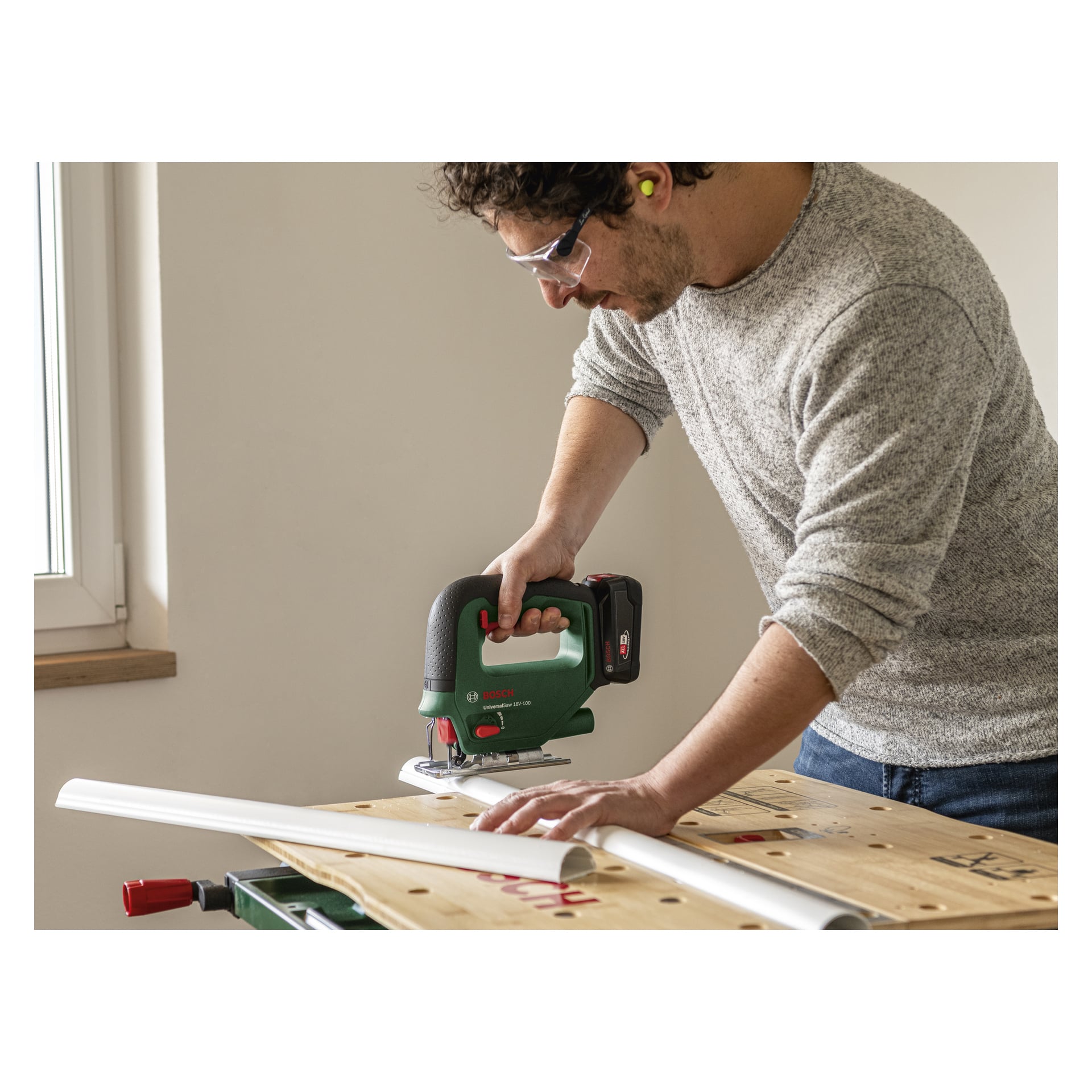 En person som bär skyddsglasögon och öronproppar använder en grön Bosch Power 4 All UniversalSaw 18V-100 sladdlös sticksåg för att skära ut en vit plast- eller PVC-list på en träarbetsbänk placerad nära ett fönster i ett rum med ljusa väggar.