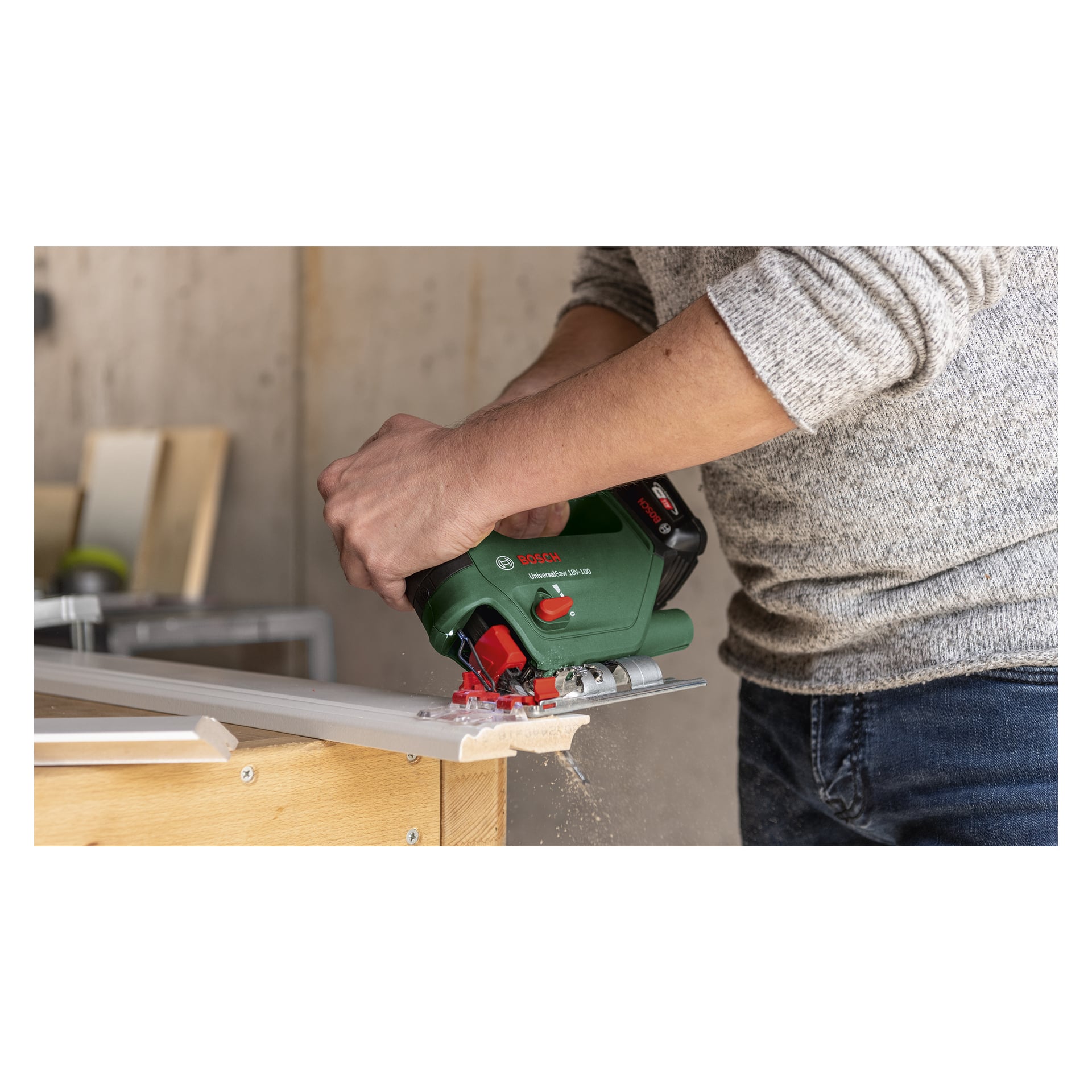 Någon i grå tröja och jeans kör Bosch Power 4 All UniversalSaw 18V-100 sladdlösa sticksåg, och skär genom en vit träskiva på en arbetsbänk medan träspån sprids från snittet.