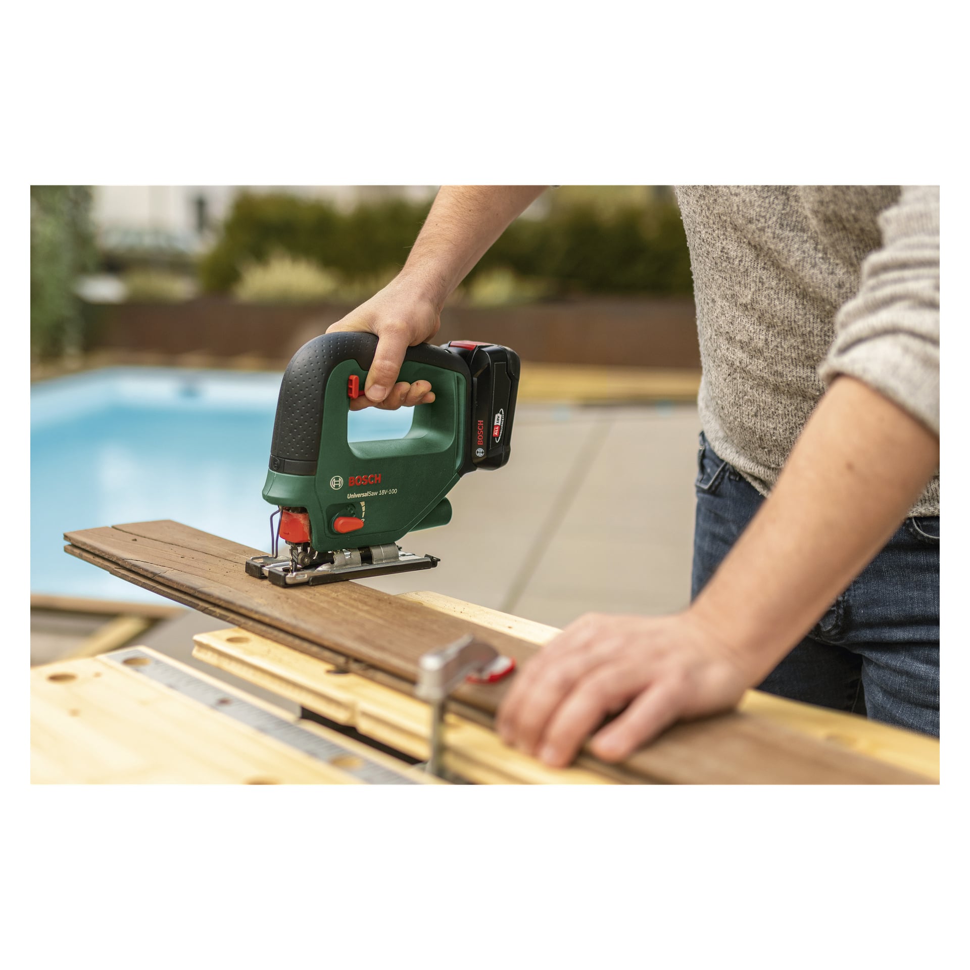 Någon använder en Bosch Power 4 All UniversalSaw 18V-100 sladdlös sticksåg för att såga igenom en träplanka på en arbetsbänk utanför vid en pool, med ansiktet utom synhåll.