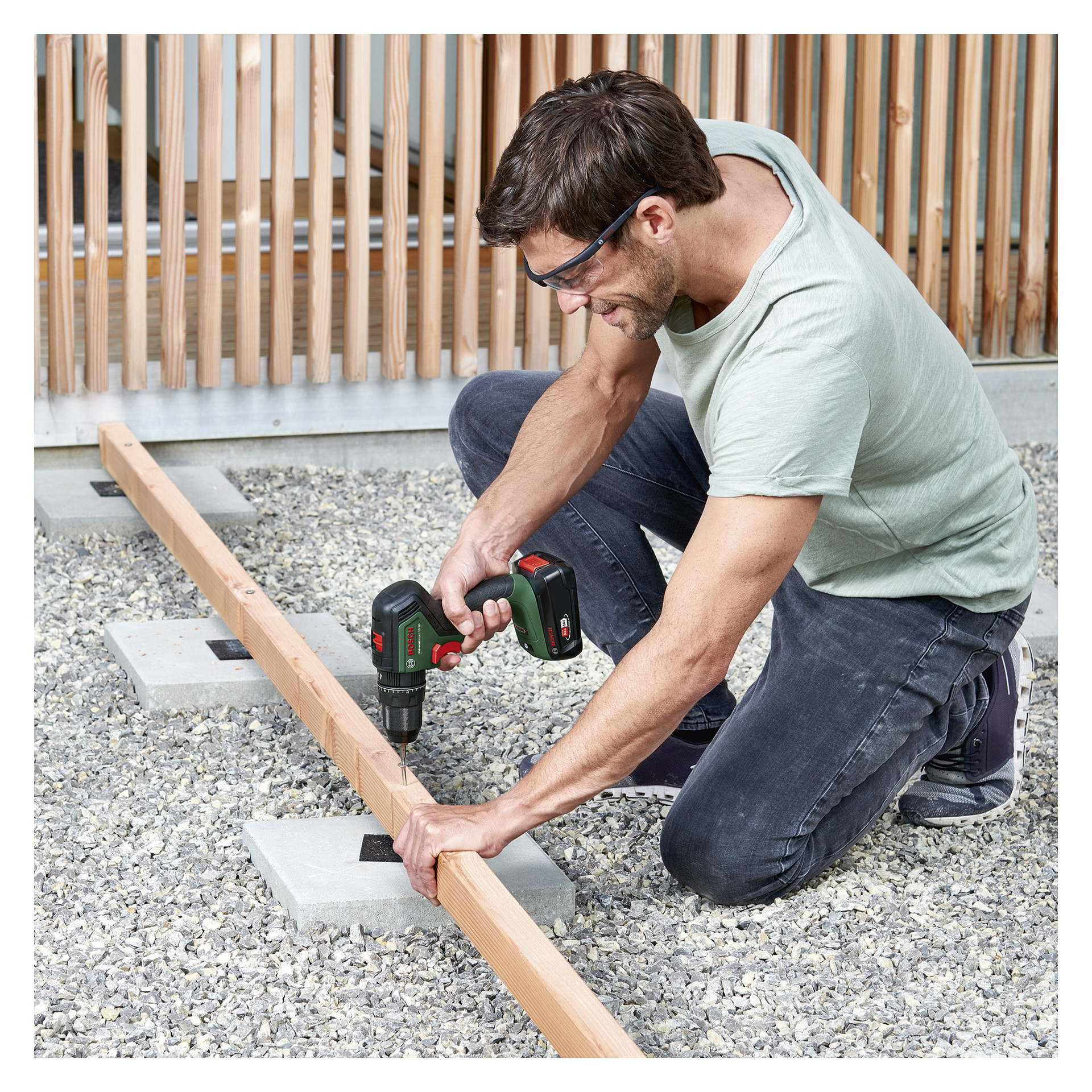 En man med skyddsglasögon knäböjer på gruset och använder en Bosch Power 4 All Brushless Impact Screwdriver Universal Impact 18V-60 för att borra i en träbalk placerad ovanpå betongblock.