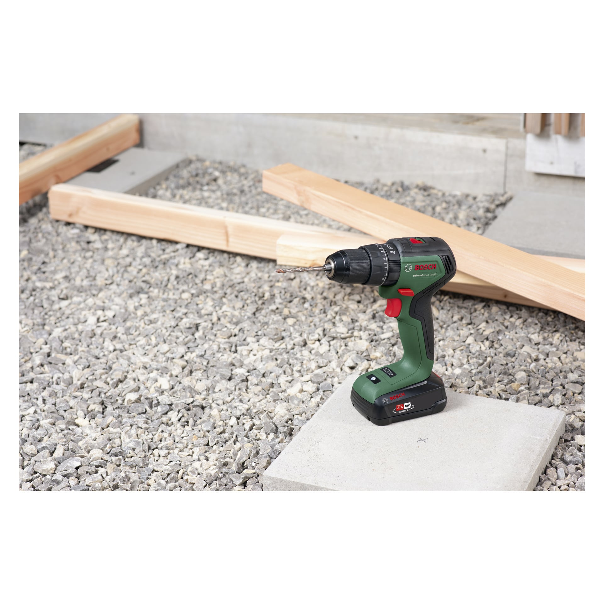 En Bosch Power 4 All Brushless Universal Impact 18V-60 skruvmejsel med borr är placerad upprätt på en betongyta, omgiven av träbalkar och grus.