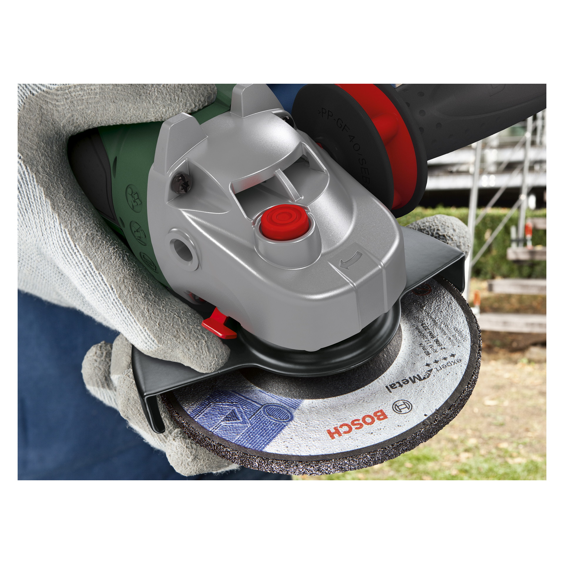 Osoba w białych rękawiczkach trzyma szlifierkę kątową Bosch Power 4 All AdvancedGrind 18V-80 zasilaną bateriami z metalowym talerzem, przygotowując się do użycia na zewnątrz. Narzędzie jest oglądane z podwyższonej, skośnej perspektywy.