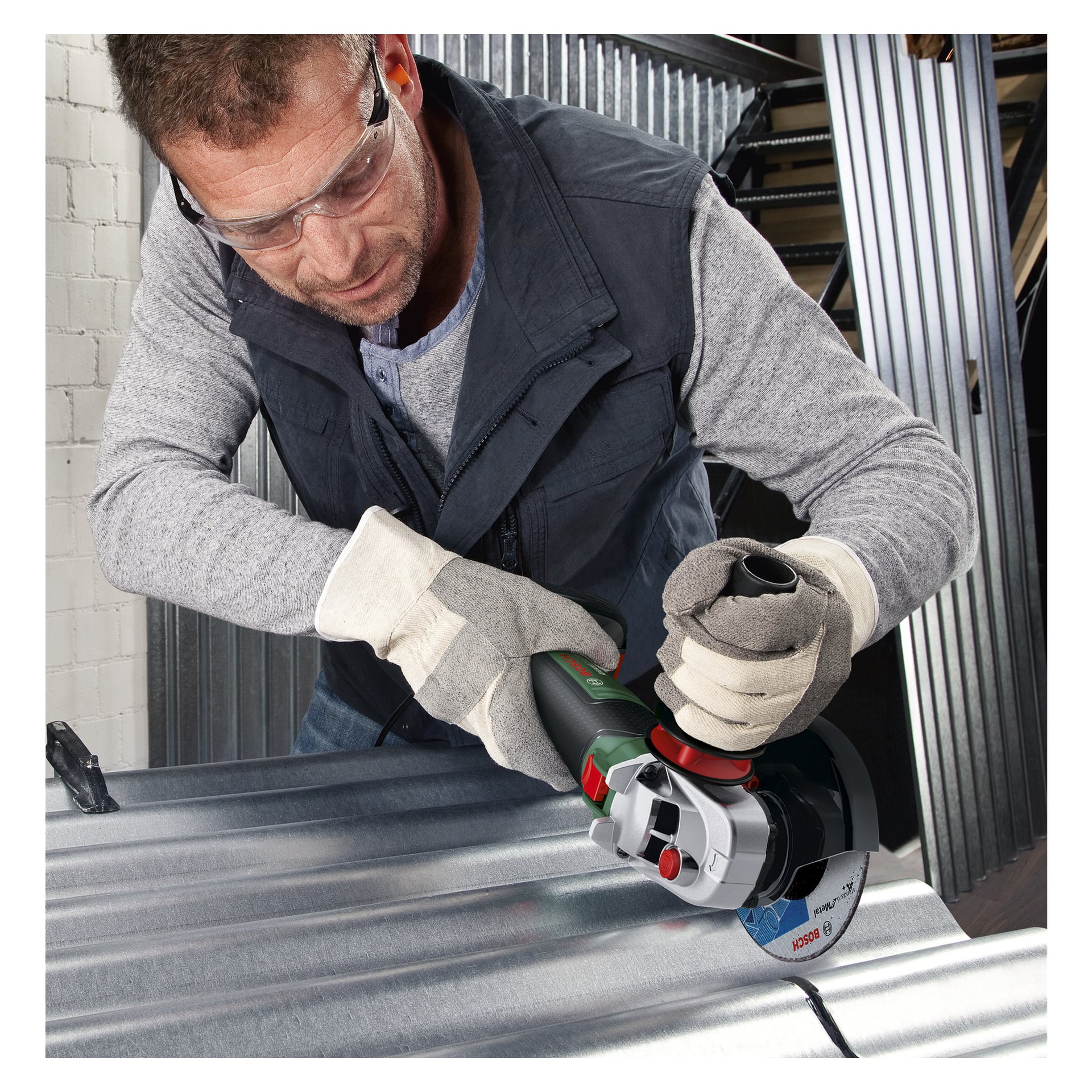 Osoba wyposażona w okulary ochronne i rękawice obsługuje bezprzewodową szlifierkę kątową Bosch Power 4 All AdvancedGrind 18V-80, aby przeciąć blachę falistą w środku, a w pobliżu znajduje się kilka stosów blach.