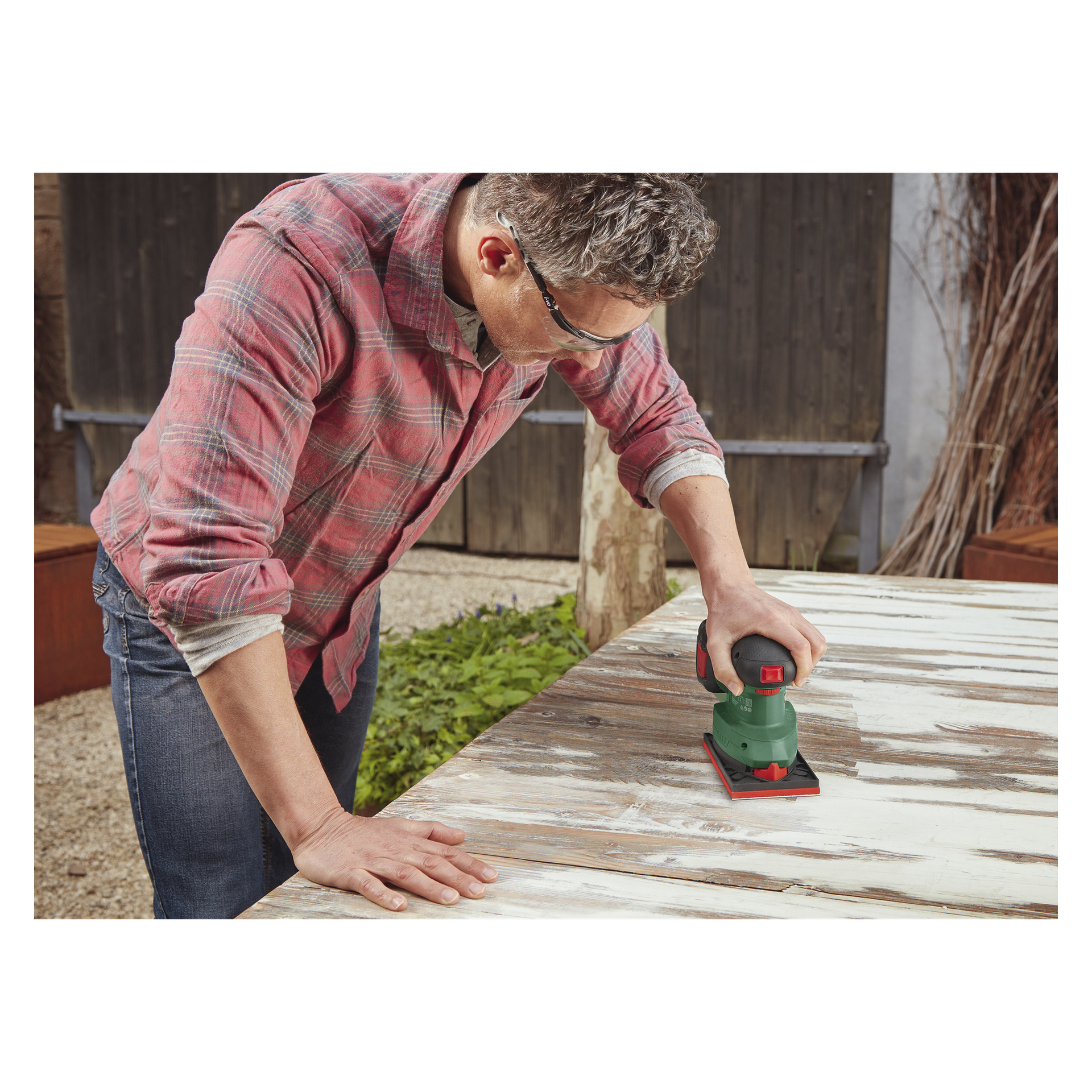 Med skyddsglasögon och rutig skjorta använder man Bosch Power 4 All UniversalSander 18V-10 sladdlös multislip för att slipa ett stort träbord utomhus.