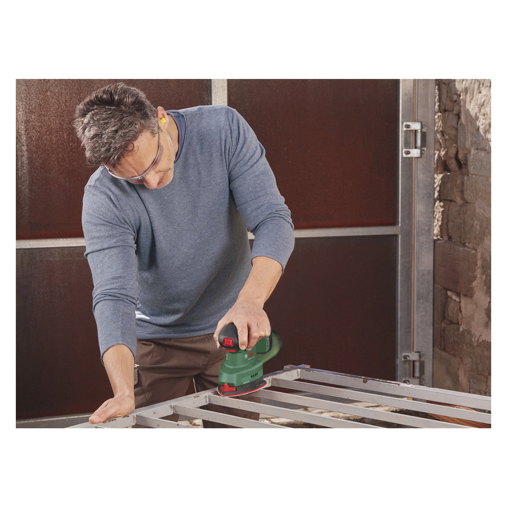 Med skyddsglasögon och hörselskydd använder man en Bosch Power 4 All UniversalSander 18V-10, där man slipar en metallram i en verkstad med väggar i sten och metall.