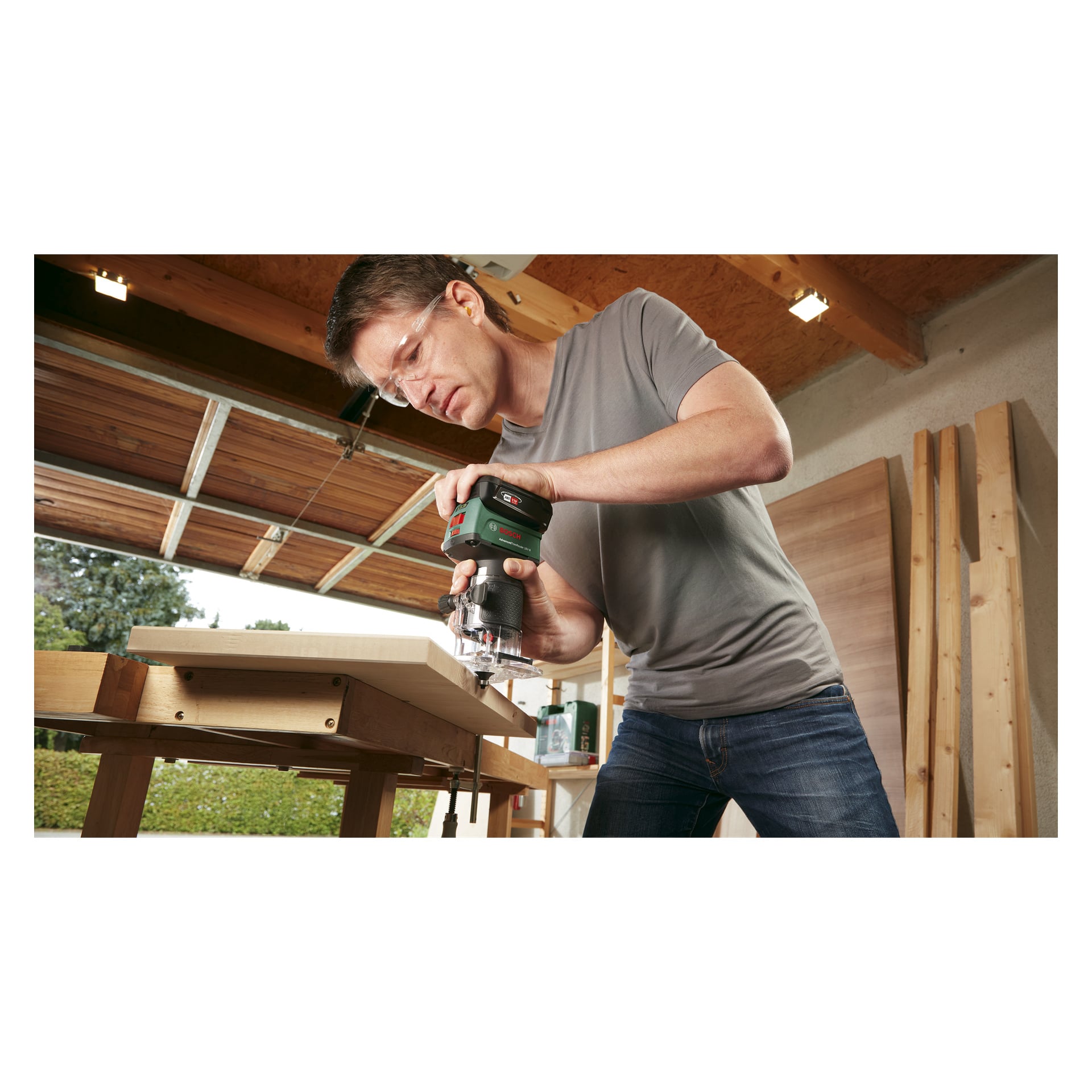En person med glasögon använder Bosch Power 4 All Router AdvancedTrimRouter 18V-8, där han skär en träplanka på en arbetsbänk i ett garage med dörren öppen.