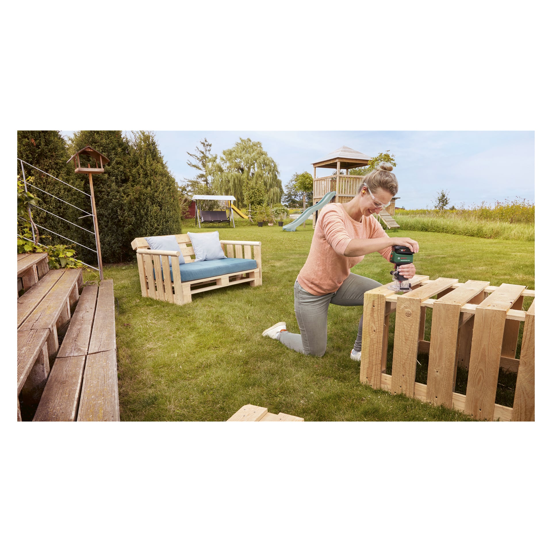 Någon knäböjer på gräset och kör en Bosch Power 4 All AdvancedTrimRouter 18V-8 på en träpall. I utomhusscenen finns möbler i trä och en lekplats, tillsammans med trappsteg och gröna växter i bakgrunden.