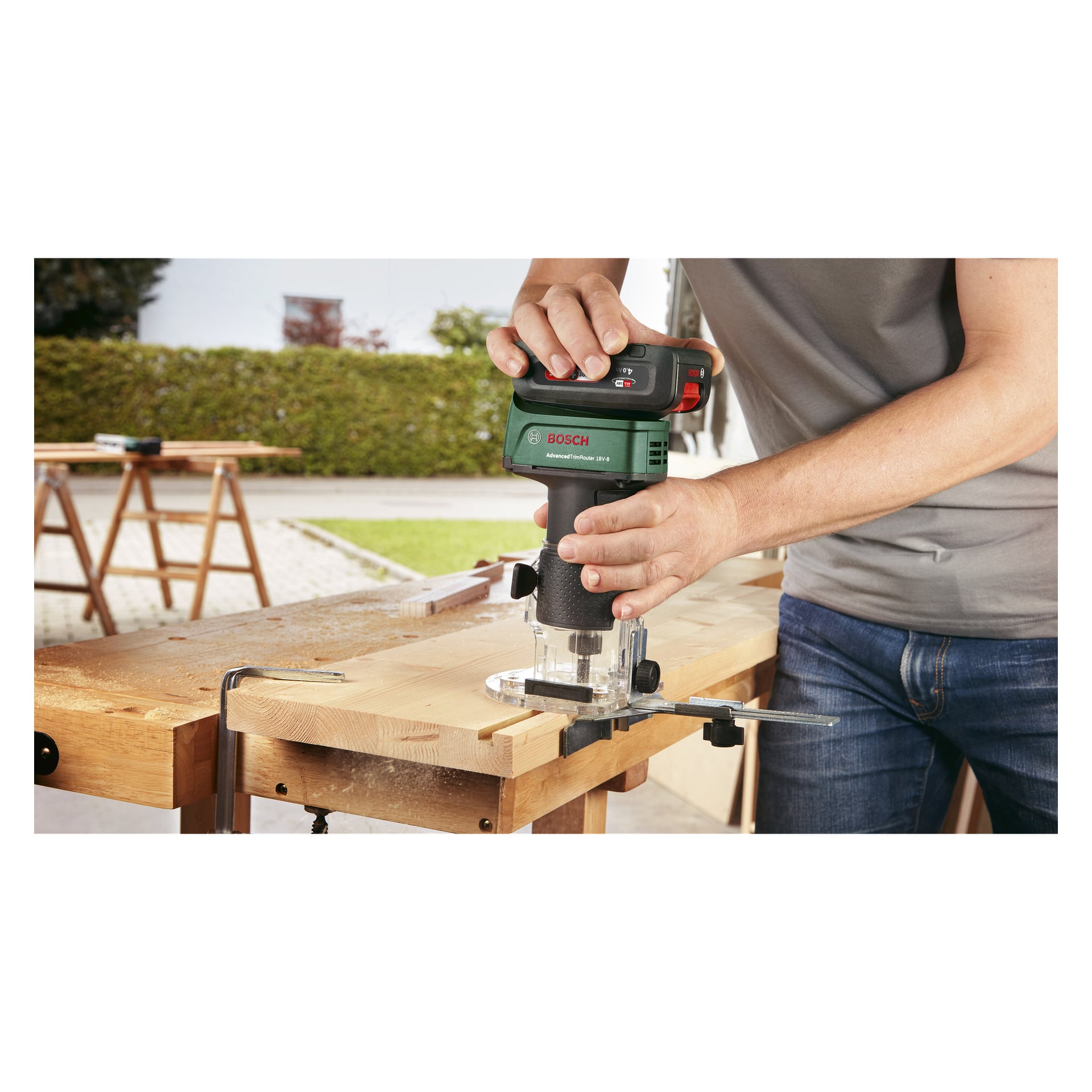 Man använder en Bosch Power 4 All Router Advanced TrimRouter 18V-8 för att forma kanten på en träskiva fäst vid en utomhusarbetsbänk, med träspån utspridda över ytan.