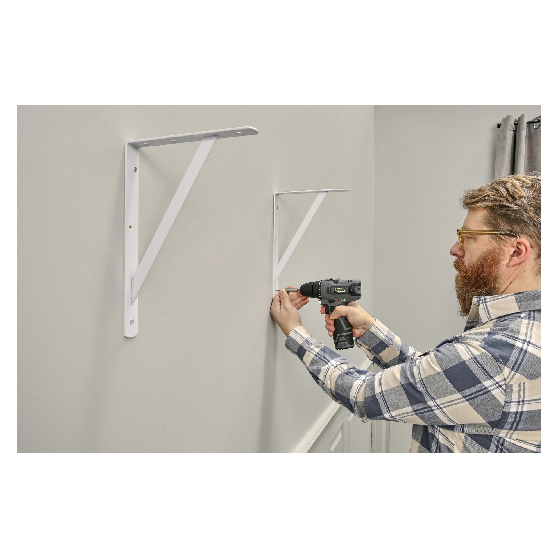 En person med glasögon och rutig skjorta använder en borrmaskin för att montera ett vitt Hard Head Reinforced fäste på en grå vägg, bredvid ett redan installerat fäste, inne i ett hus.