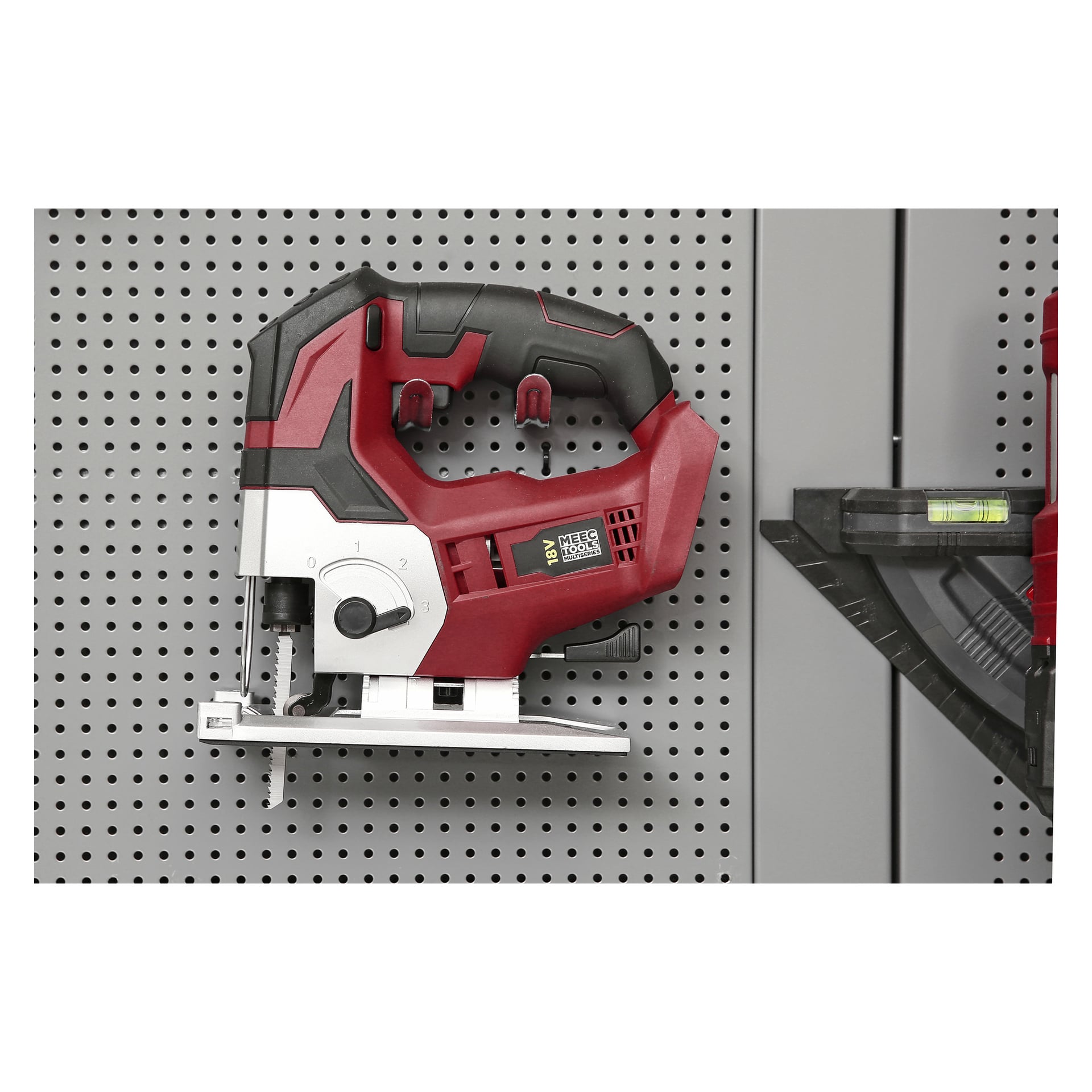 En röd och svart Meec Tools Multiseries Jigsaw JS-18V HOME 300 är fäst vid en grå perforerad metallpegboard. På höger sida kan man se en del av ett svart mätverktyg och ett grönt vattenpass med bubbla.