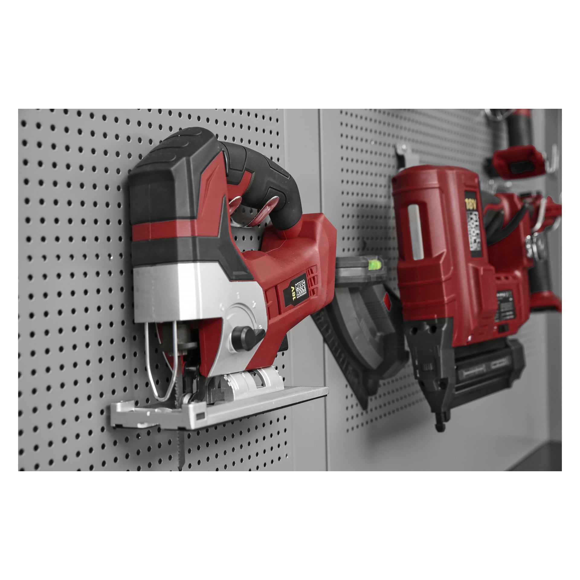 En närbild av en Meec Tools Multiseries Jigsaw JS-18V HOME 300 och en spikpistol som visas på en grå perforerad pinnvägg i en verkstad eller verktygsförvaring.