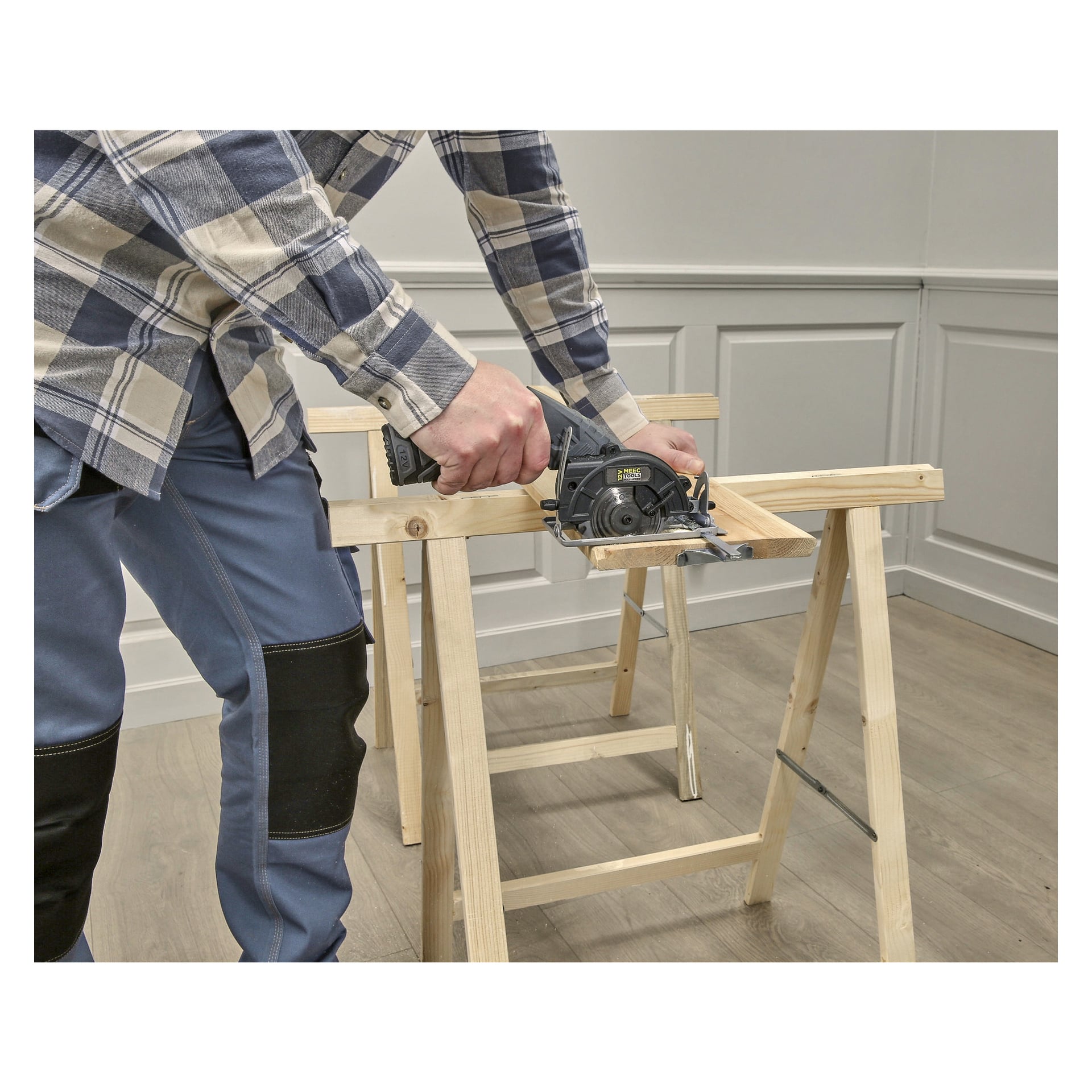 Iklädd en rutig skjorta och blå byxor använder man Meec Tools Multiseries 12 V Compact cirkelsåg för att såga en träplanka på såghästar i ett starkt upplyst rum med panelpaneler och ljusa trägolv.