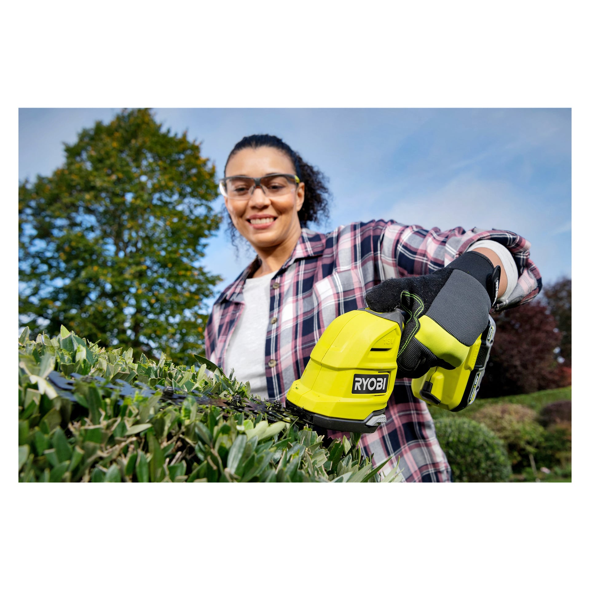 Med skyddsglasögon och handskar använder man Ryobi One+ Multi-trimmer RY18GSA-0 för att klippa en häck utomhus, med träd och blå himmel i bakgrunden.