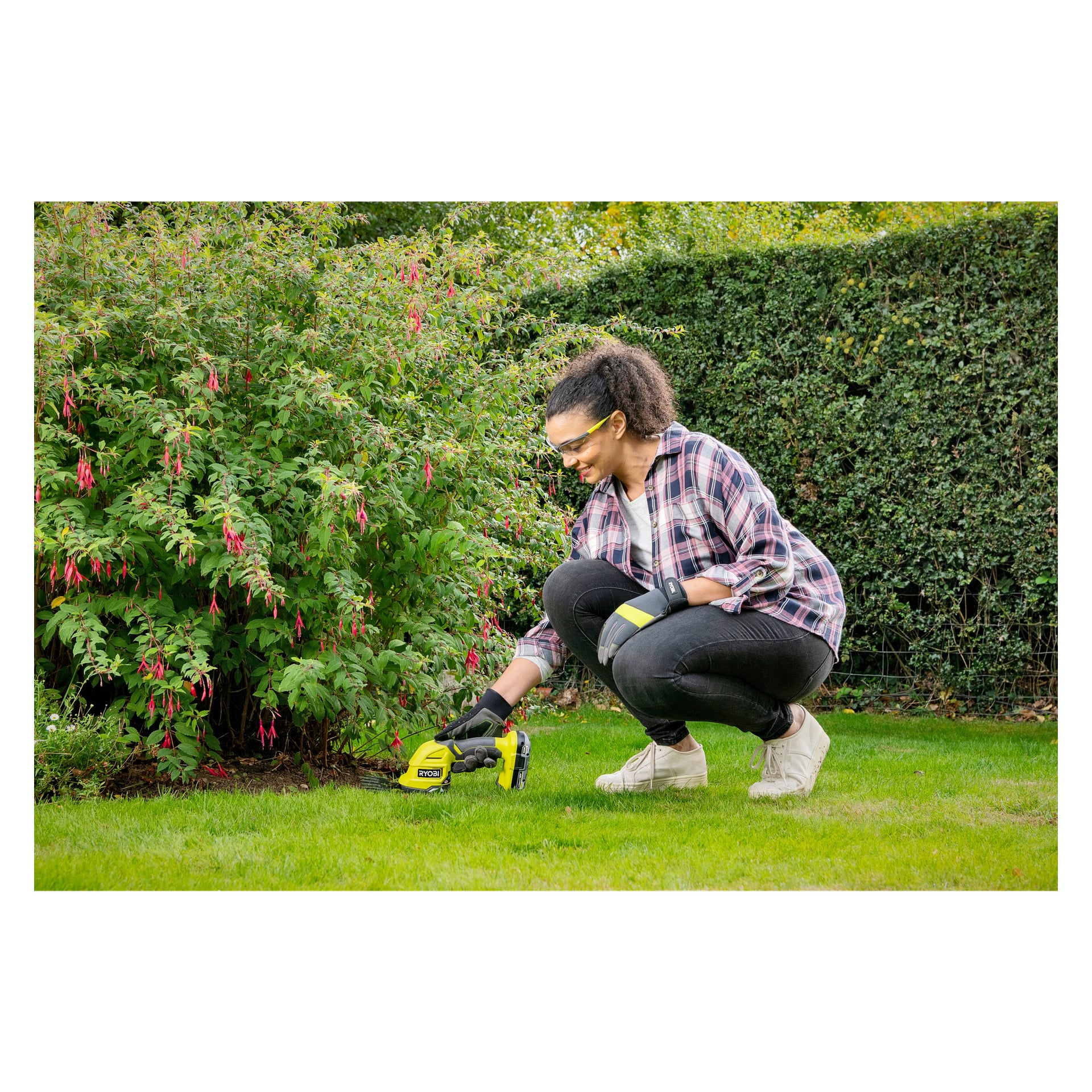 En kvinna med skyddsglasögon och handskar sitter hukande på gräset och trimmar kanten av en rabatt med en gul Ryobi One+ Multi-trimmer RY18GSA-0 sladdlös verktyg. I bakgrunden finns en stor buske med rosa blommor och en hög grön häck.