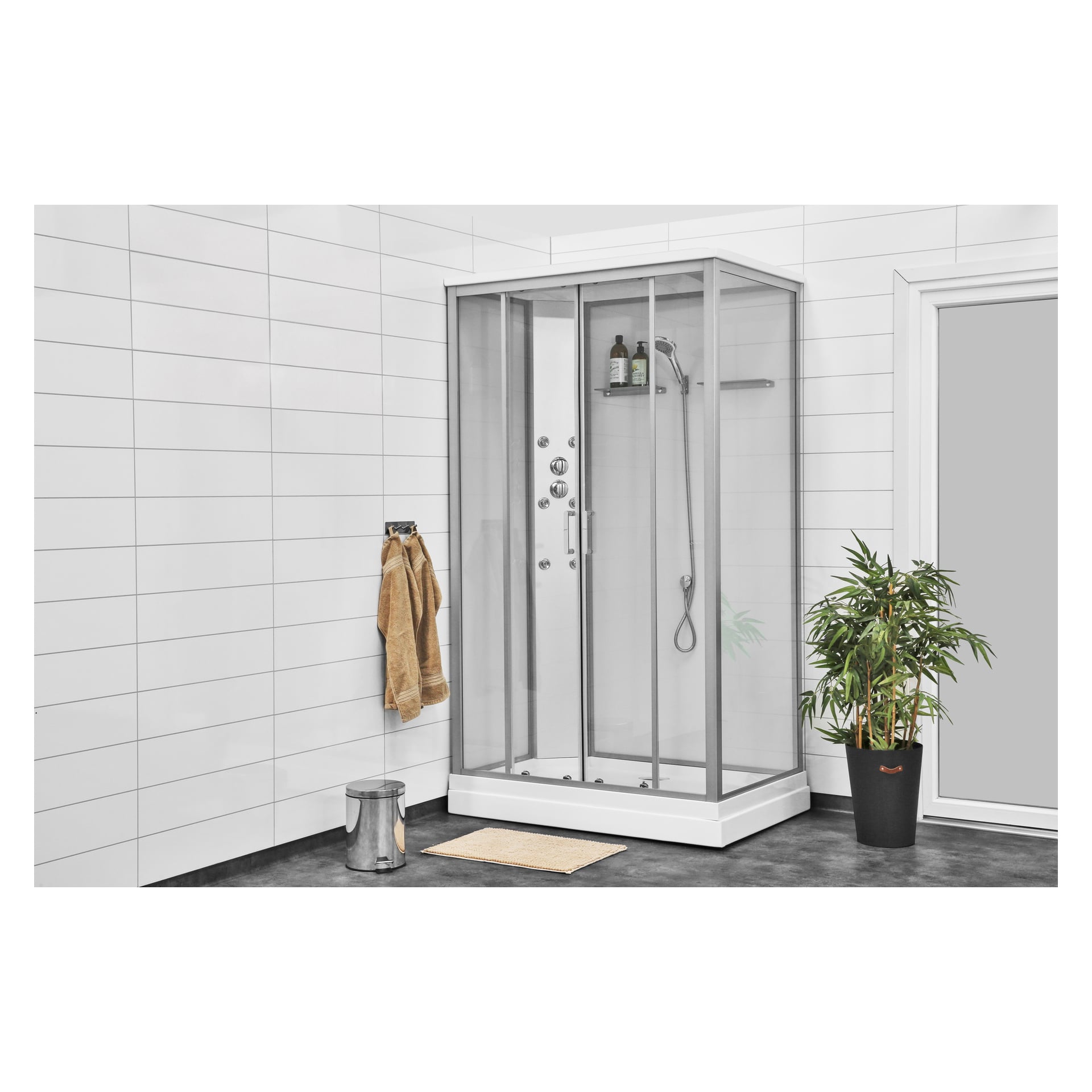 Et moderne bad med EKVIP dusjkabinett og takdusj i hjørnet, et håndkle på krok i nærheten, sølvsøppelbøtte, beige matte, potteplante og hvite flislagte vegger.