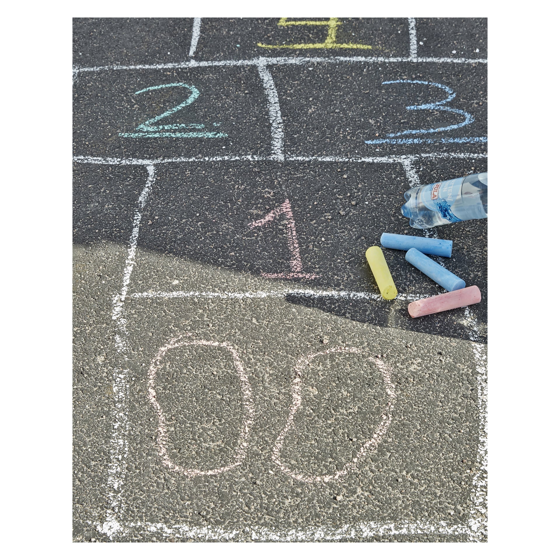Et hoppeskotsmønster nummerert 1 til 5 er tegnet på asfalt i gult, rosa og blått ved bruk av Kayobas Street Chalks 20-pakke. Noen fargerike krittpinner fra samlingen og en klar plastflaske ligger spredt på gulvet i nærheten.