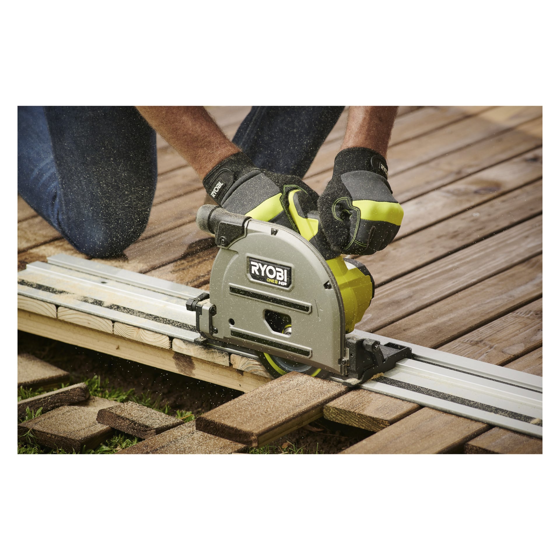 En person med handske använder en batteridriven Ryobi One+ HP dyksåg med styrskena (RPLS18X-0) för att såga en träplanka på en utomhusaltan, omgiven av sågspån nära skärpunkten.