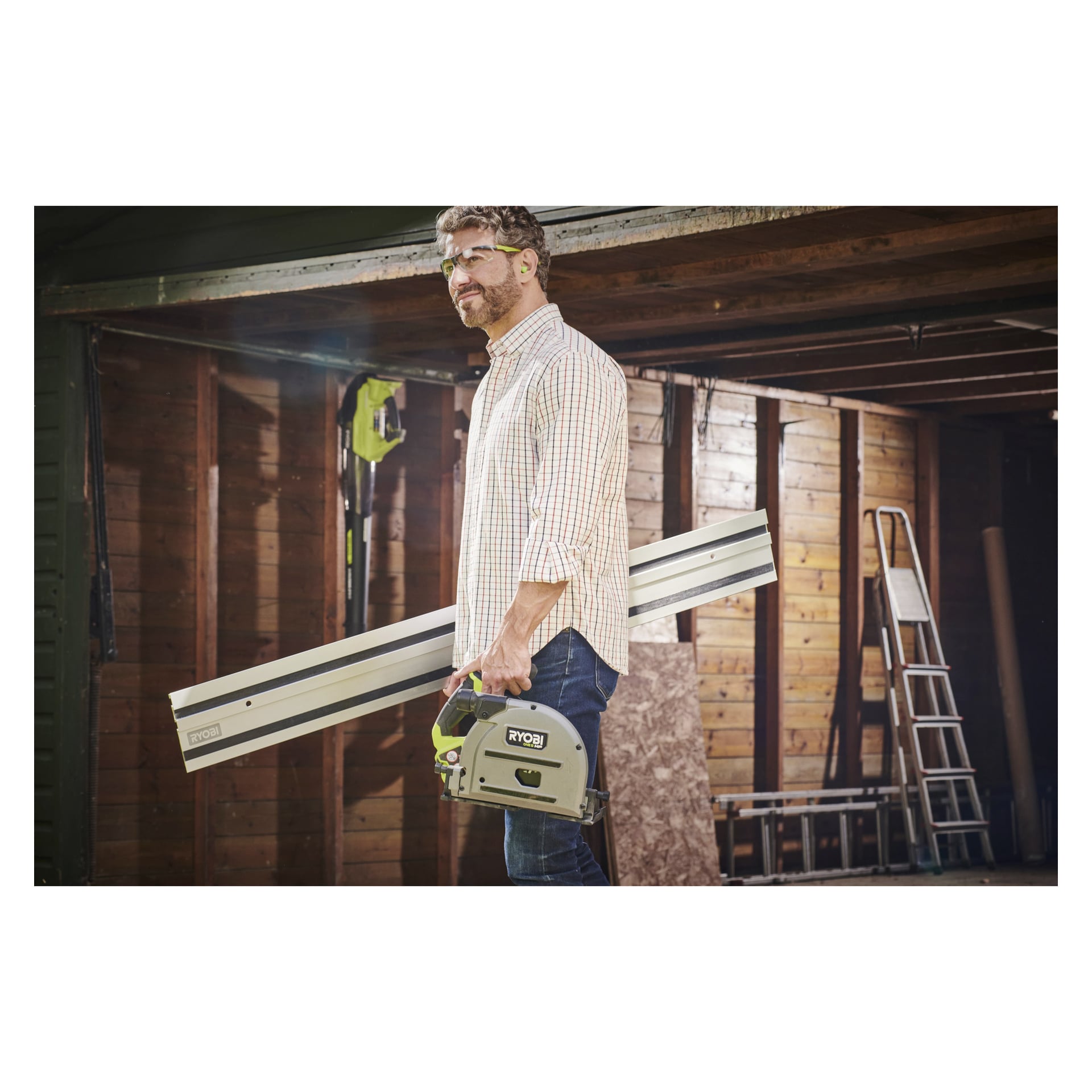 En person med skyddsglasögon håller i en Ryobi One+ HP batteridriven dyksåg och två långa Ryobi-styrskena (RPLS18X-0), och står utanför ett trägarage där stegar och byggmaterial syns.