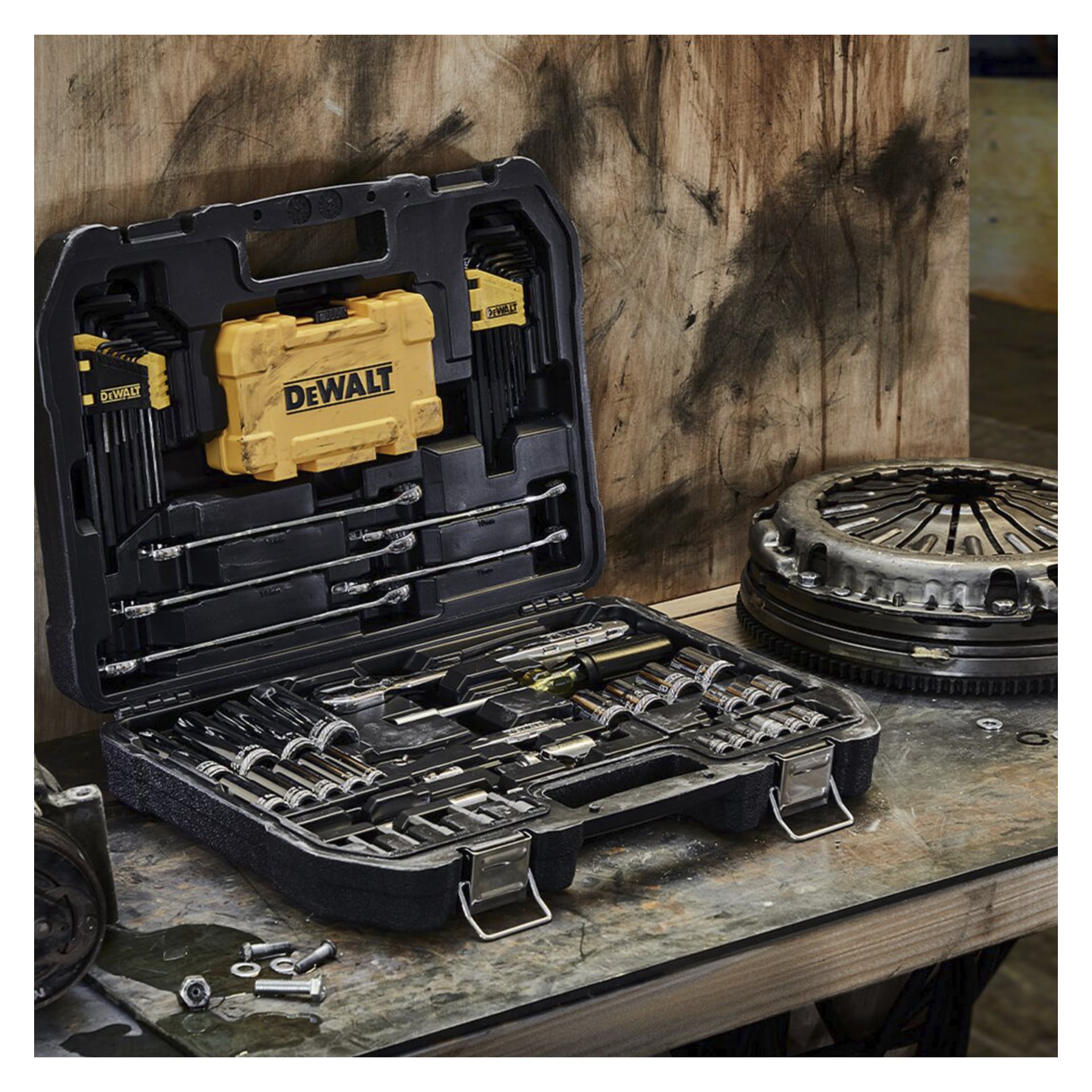 Ett svart DeWalt-skiftnyckelset med 89 delar står öppet på en arbetsbänk och avslöjar olika hylsor, skiftnycklar och bitar som är prydligt arrangerade inuti. En gul DeWalt-låda placeras också i fodralet. Bakgrunden visar bildelsar och träytor.
