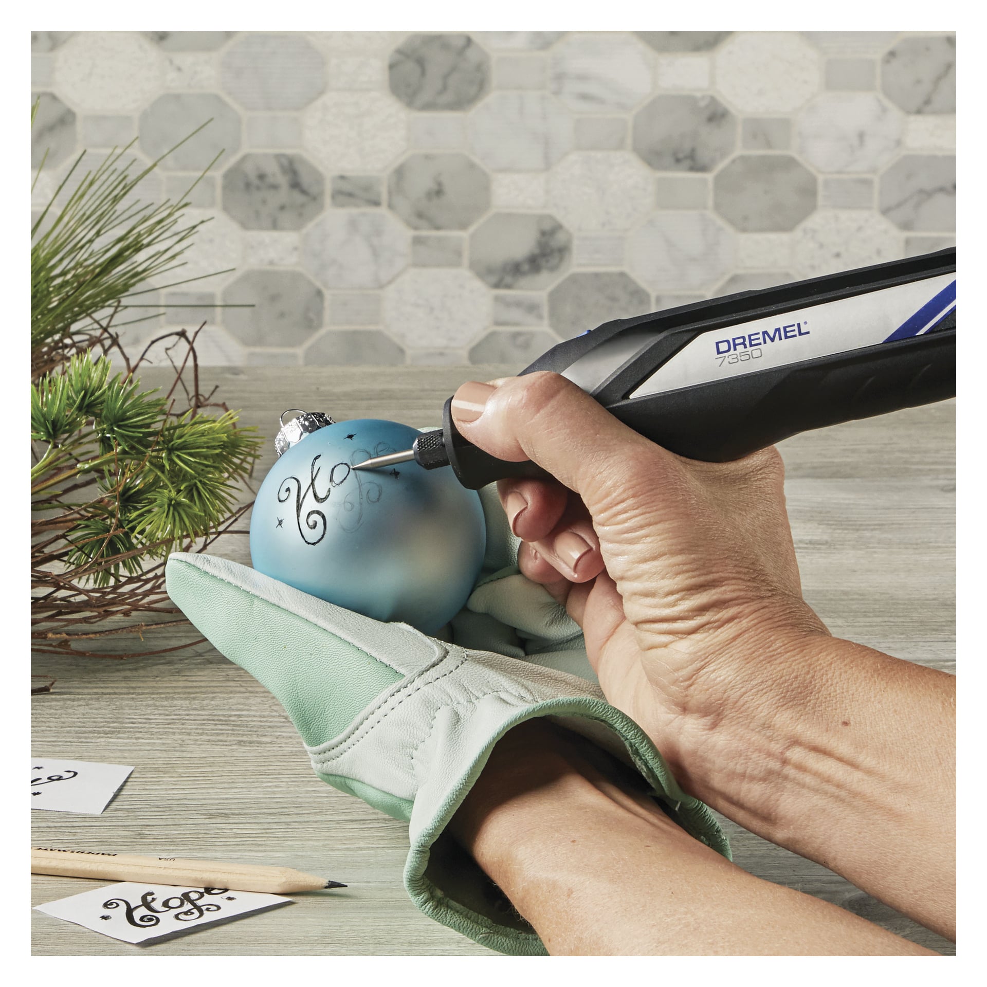 En person använder en Dremel-gravyrnål 105 (3-pack) för att snida dekorativ text på en blå julgransprydnad, och håller den med en handske nära tallgrenar på en lätt träyta.