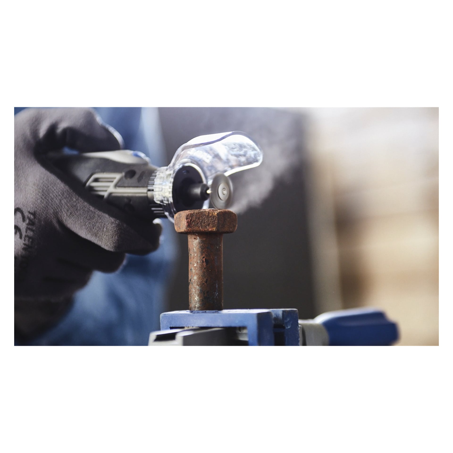 Handskbeklädda händer använder en Dremel Steel-borste 428 för att rengöra rost från en metallbult som hålls hårt i ett skruvstäd, med damm och skräp synligt virvlande runt bulten under rengöringen.