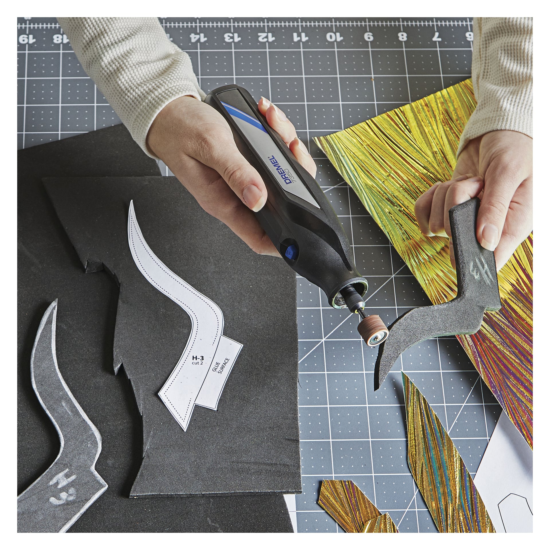 En person använder ett Dremel Sanding Belt 6 Pack för att finslipa kanten på en svart skumutskärning placerad på en rutnätsmatta, med mönstermallar, färgglada material och diverse pysselmaterial utspridda i närheten.