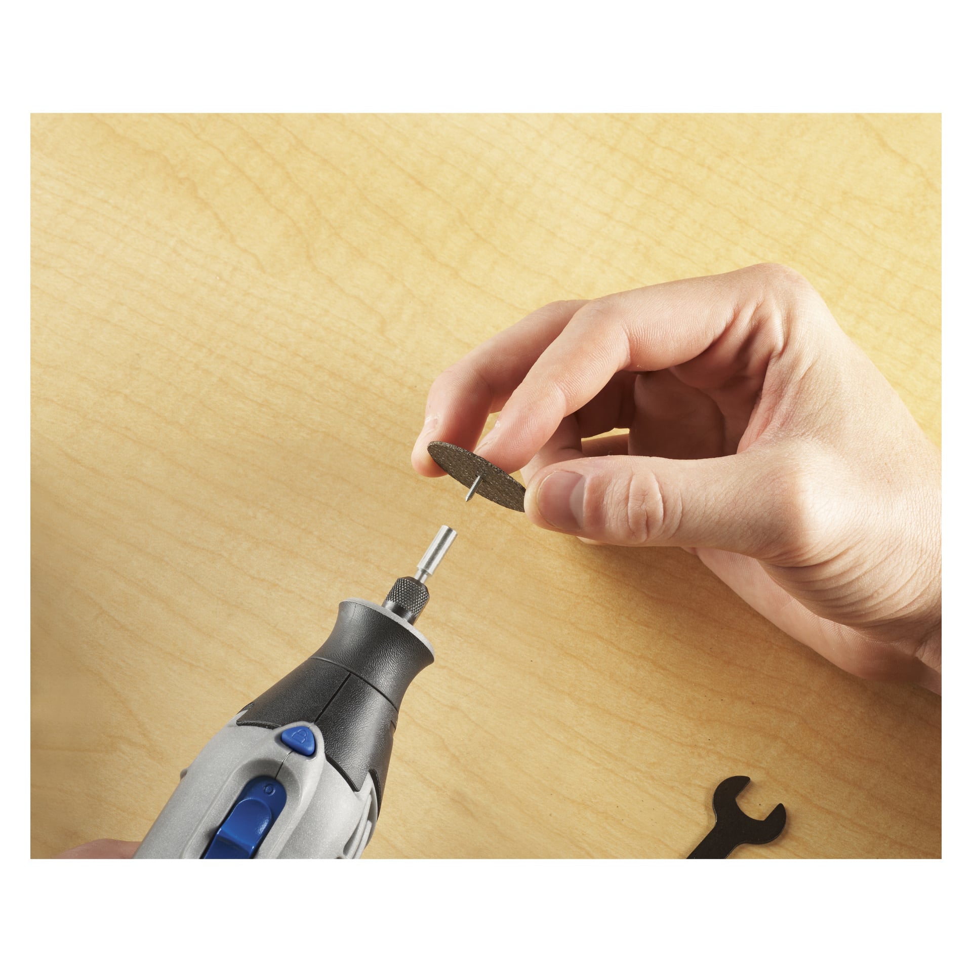 Någon fäster en cirkulär skärskiva på ett Dremel Spindle-set 402 (4-pack) roterande verktyg ovanför en lätt träyta. Den lilla svarta skiftnyckeln från setet vilar på bordet i närheten.