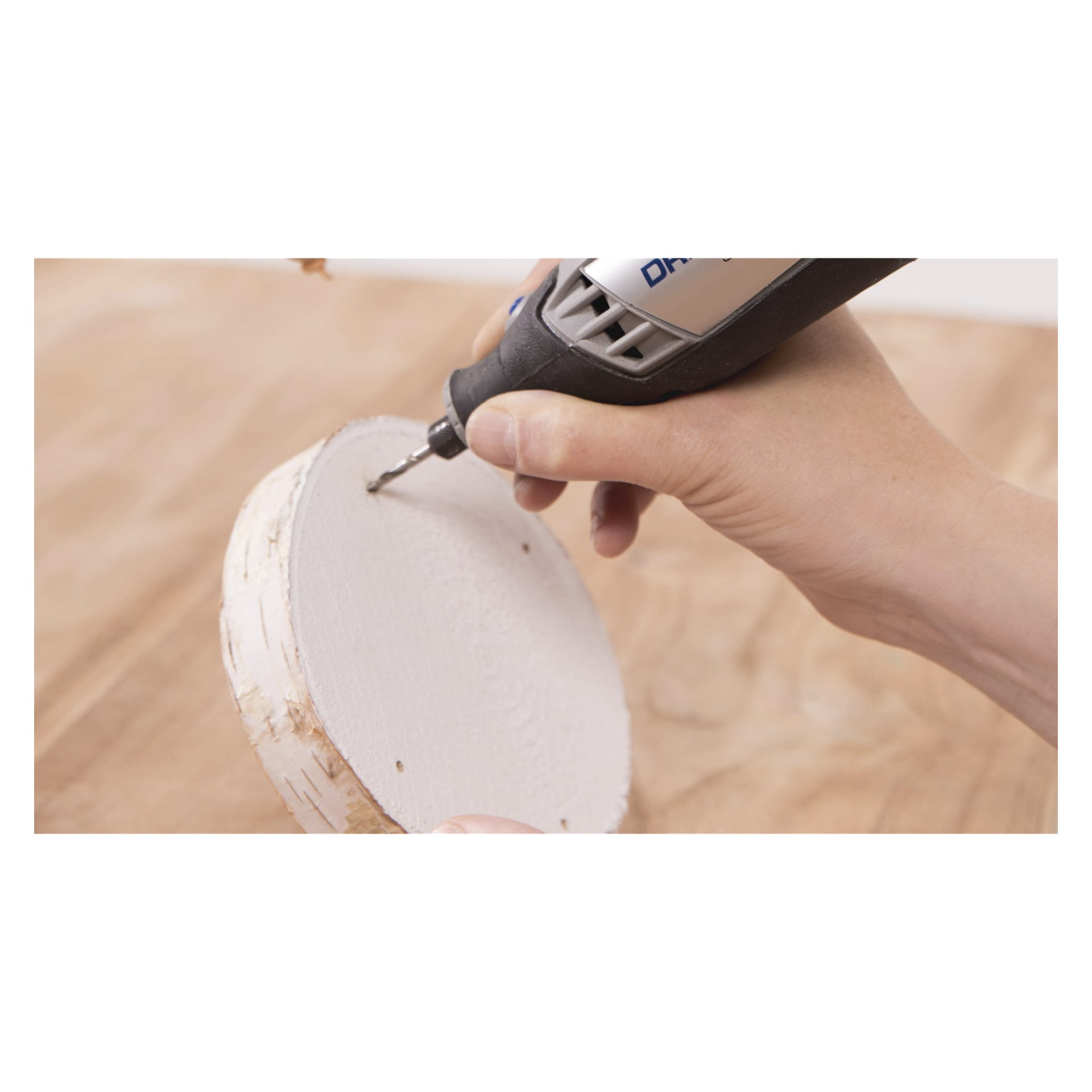 En person borrar ett hål i en rund, vitmålad träbit med hjälp av ett Dremel Woodworking set 681 roterande verktyg, håller träet med ena handen och arbetar över en träyta.