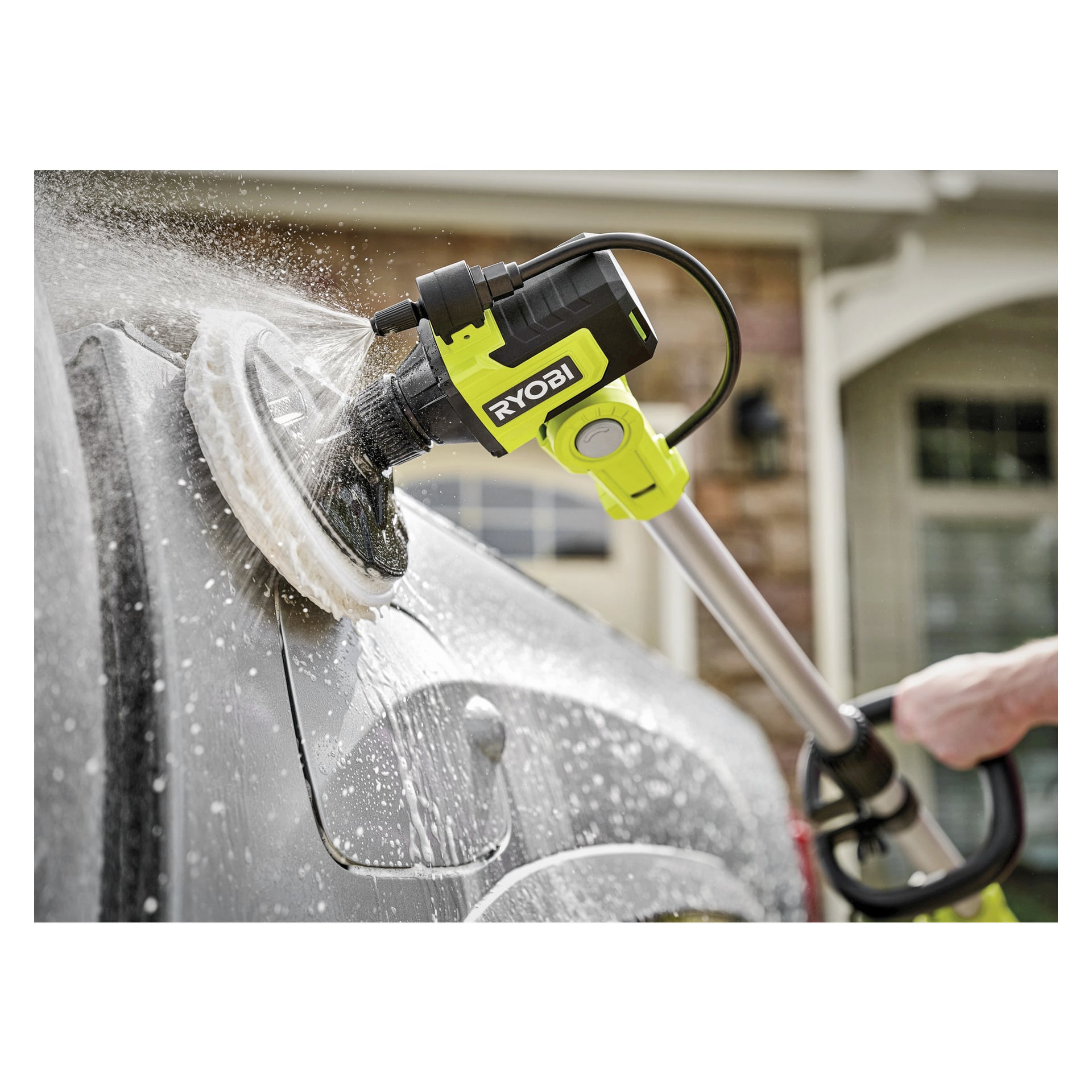 En person använder en Ryobi One+ roterande tvättborste RWTS18-0 utrustad med en skumdyna och sprutande vatten för att tvätta baksidan på en mörkfärgad bil utomhus, med ett hus synligt i närheten.