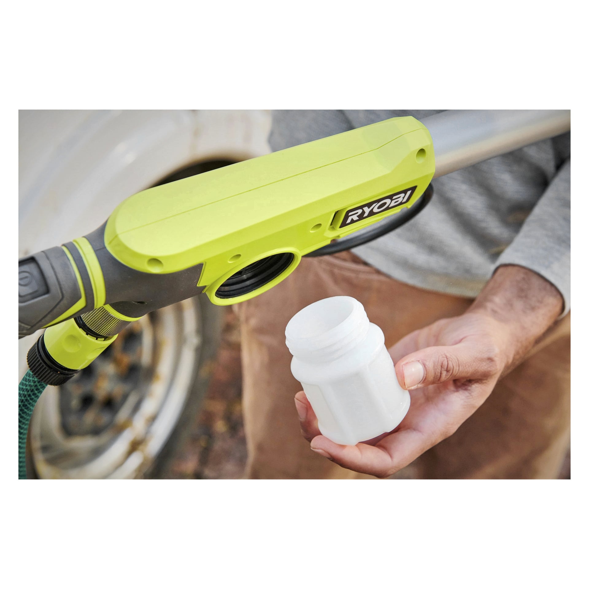 Någon håller Ryobi One+ roterande tvättborste RWTS18-0 nära munstycket på en grön Ryobi högtryckstvätt och förbereder sig för att koppla in den. Endast deras hand och bål syns, med ett bildäck synligt bakom dem.