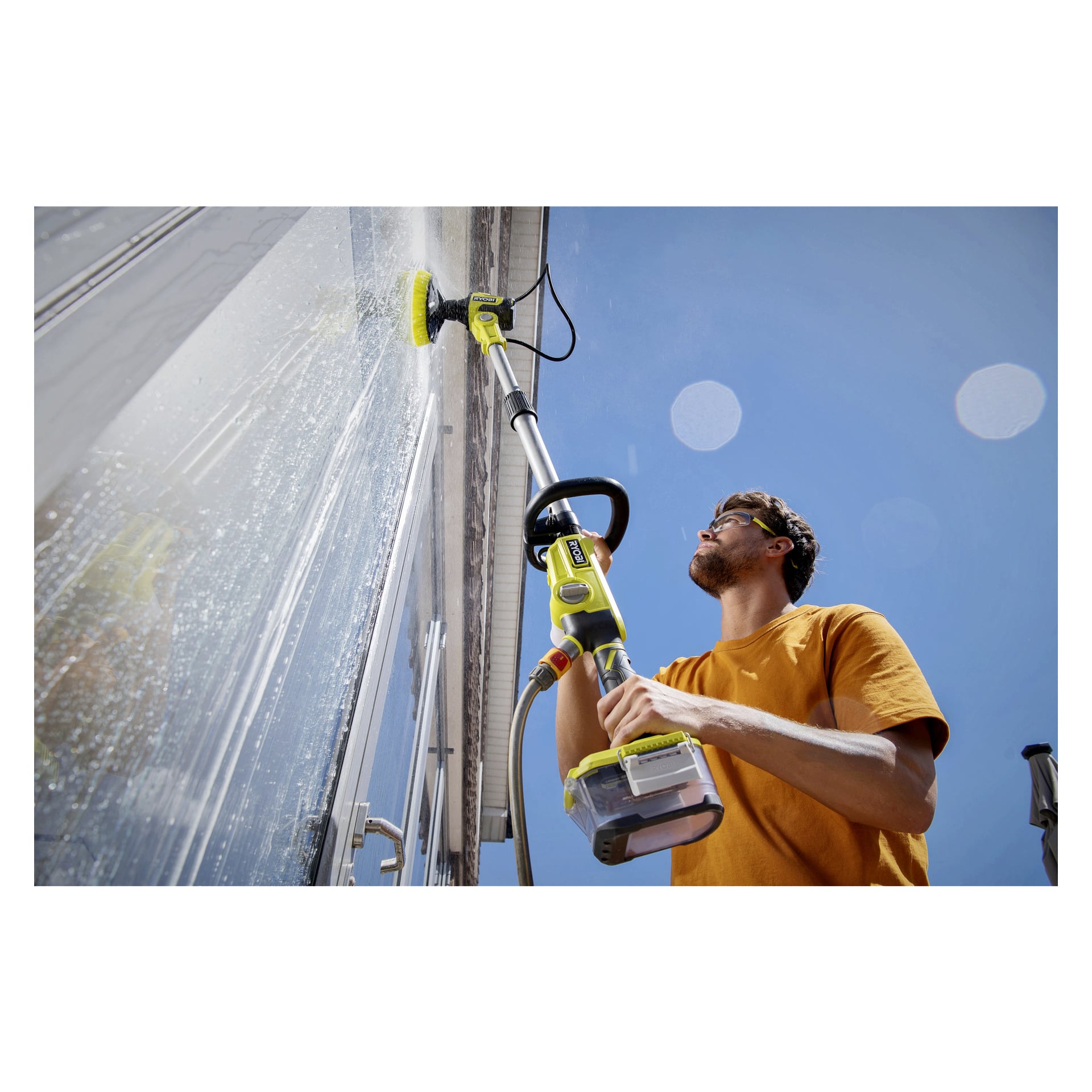 Med en orange skjorta använder man en Ryobi One+ roterande tvättborste RWTS18-0 fäst vid en förlängningsstolpe för att tvätta utsidan av ett fönster en ljus, solig dag. Fönstret visar tvålskum och vattenstrimmor.