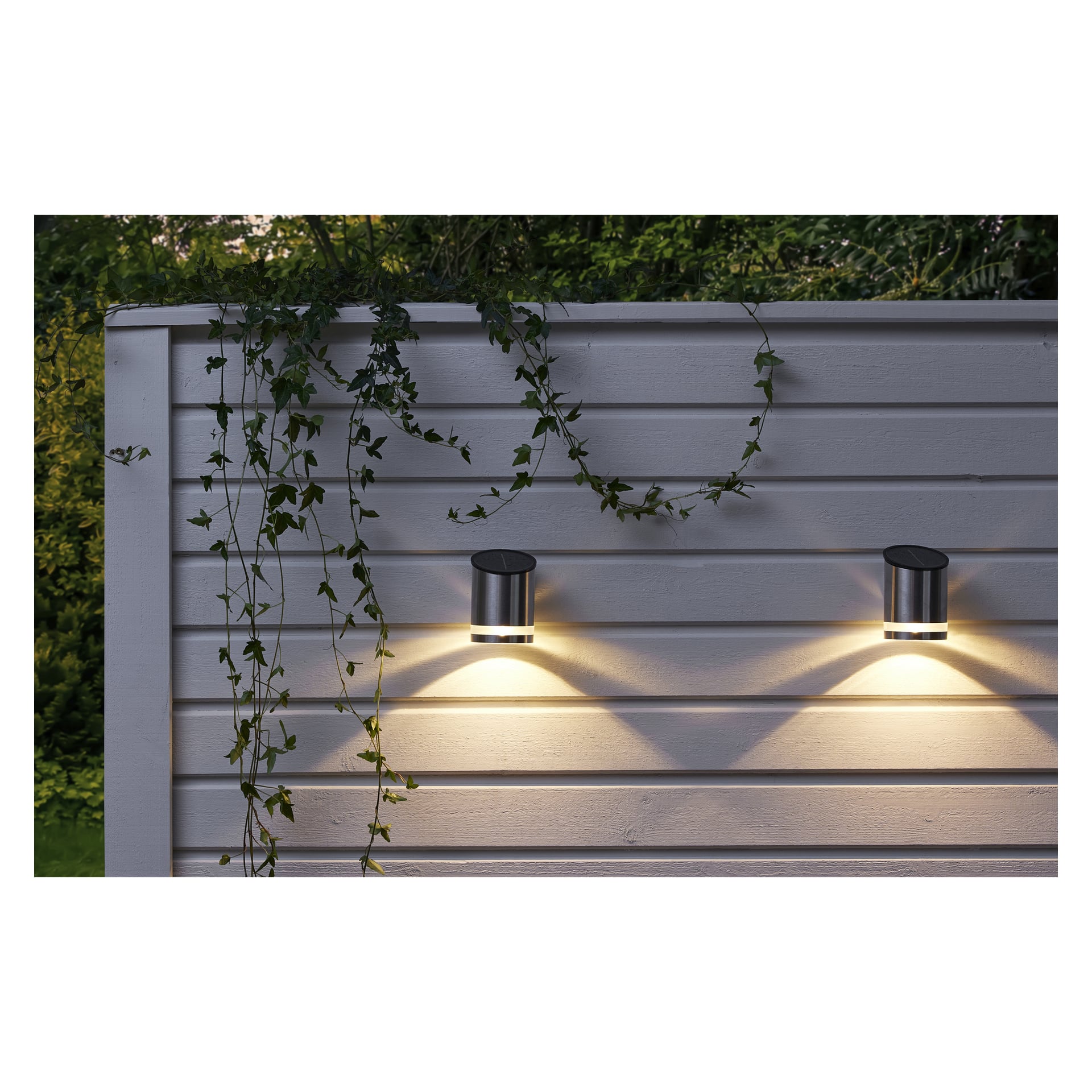 To Anslut Wall-lamper LED-lys er montert på et gjerde med grønne vinranker som kaster V-formede lysmønstre. Trær og løvverk kan sees i bakgrunnen.