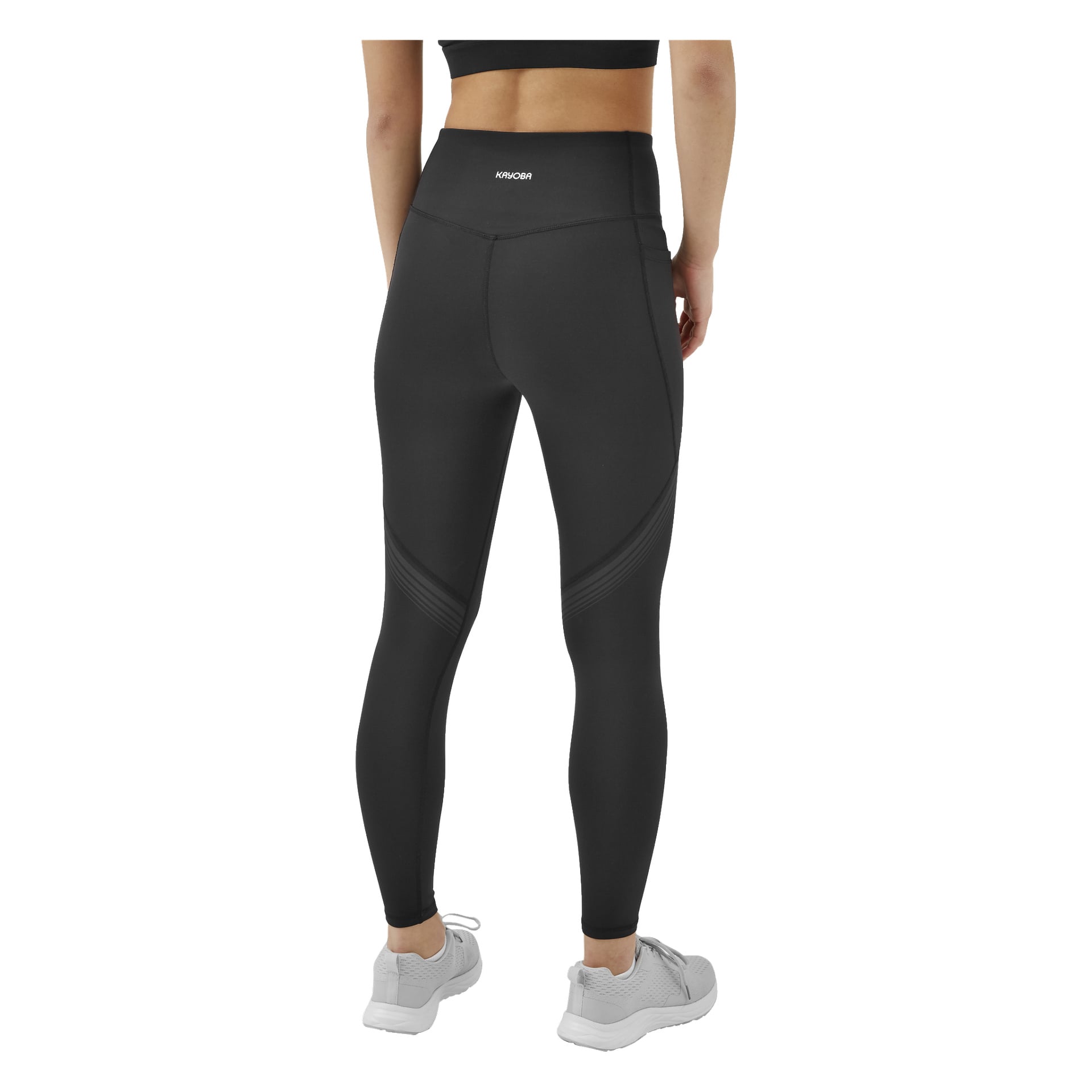 En person står med ryggen mot kameran, klädd i svarta Kayoba Tights damleggings i Boston med framträdande sömdetaljer och en midjebandslogga, tillsammans med en svart sport-bh och ljusgrå träningssneakers.
