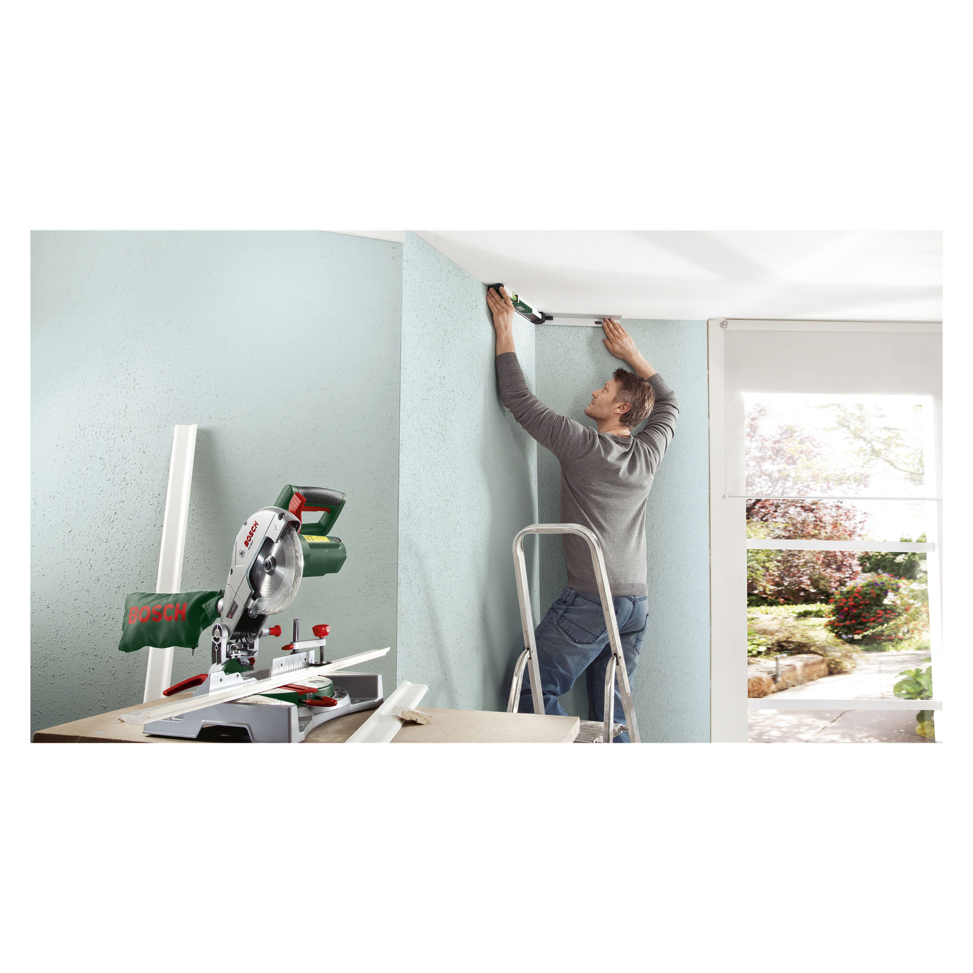 En man på en stege installerar lister längs taket på en ljusblå vägg, noggrant mätt med Bosch Digital vinkelmätare UniversalAngle. Solljus silas in genom ett stort fönster i bakgrunden, och på golvet nedanför ligger en elektrisk såg, flera listsektioner och verktyget UniversalAngle.