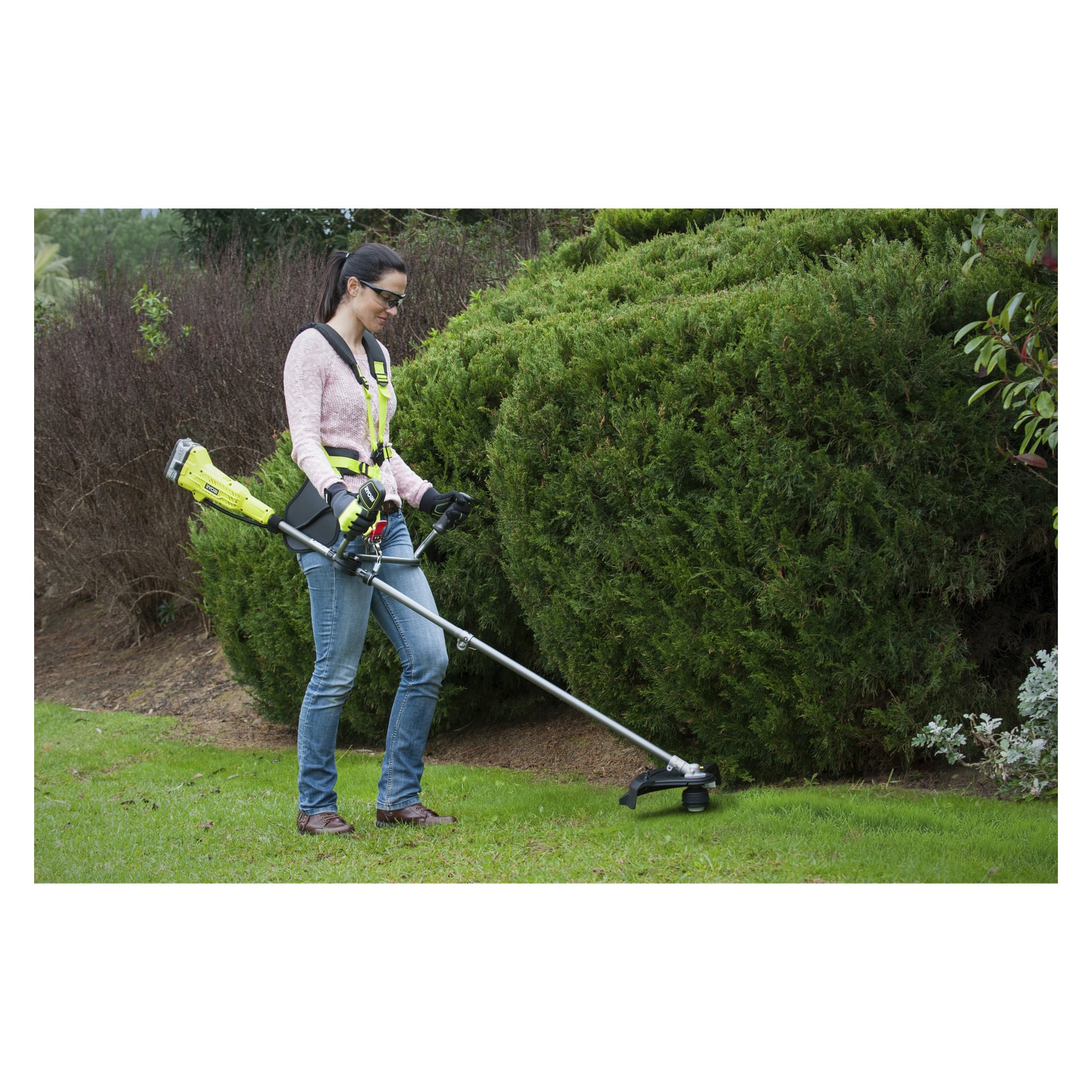 Med skyddsutrustning använder man Ryobi One+ batteridriven buskklippare OBC1820B för att klippa gräs längs en häck. De står på en grön gräsmatta, håller verktyget med båda händerna och koncentrerar sig på området de kantar.